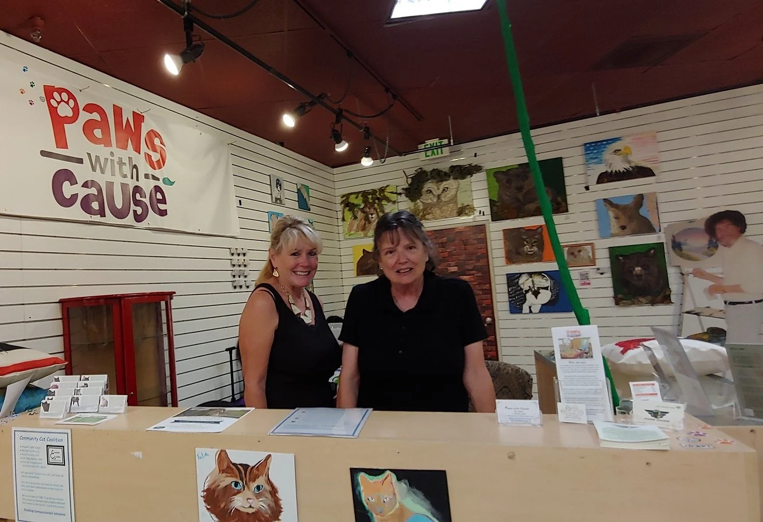 Two women smiling at a booth with art and informational materials in front. Behind them, a large sign reads 'PAWS with a purpose' and the wall is decorated with various animal portraits, including cats and owls.