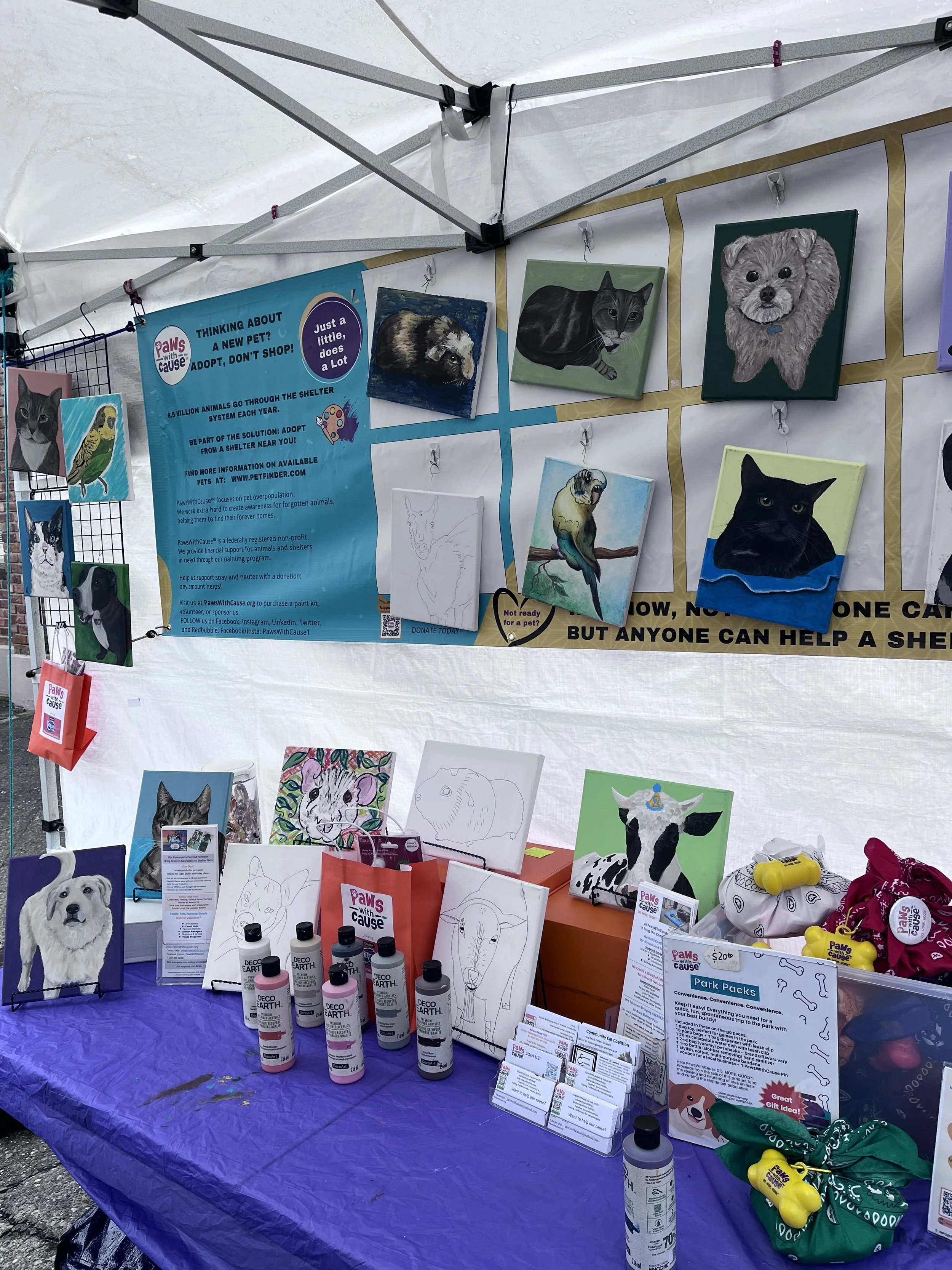 A booth at an outdoor event displays painted pet portraits, informational posters about pet adoption, and pet supplies. Paintings depict cats, dogs, and other animals, with some still being sketched. Brochures and bottled paint are arranged on a purp