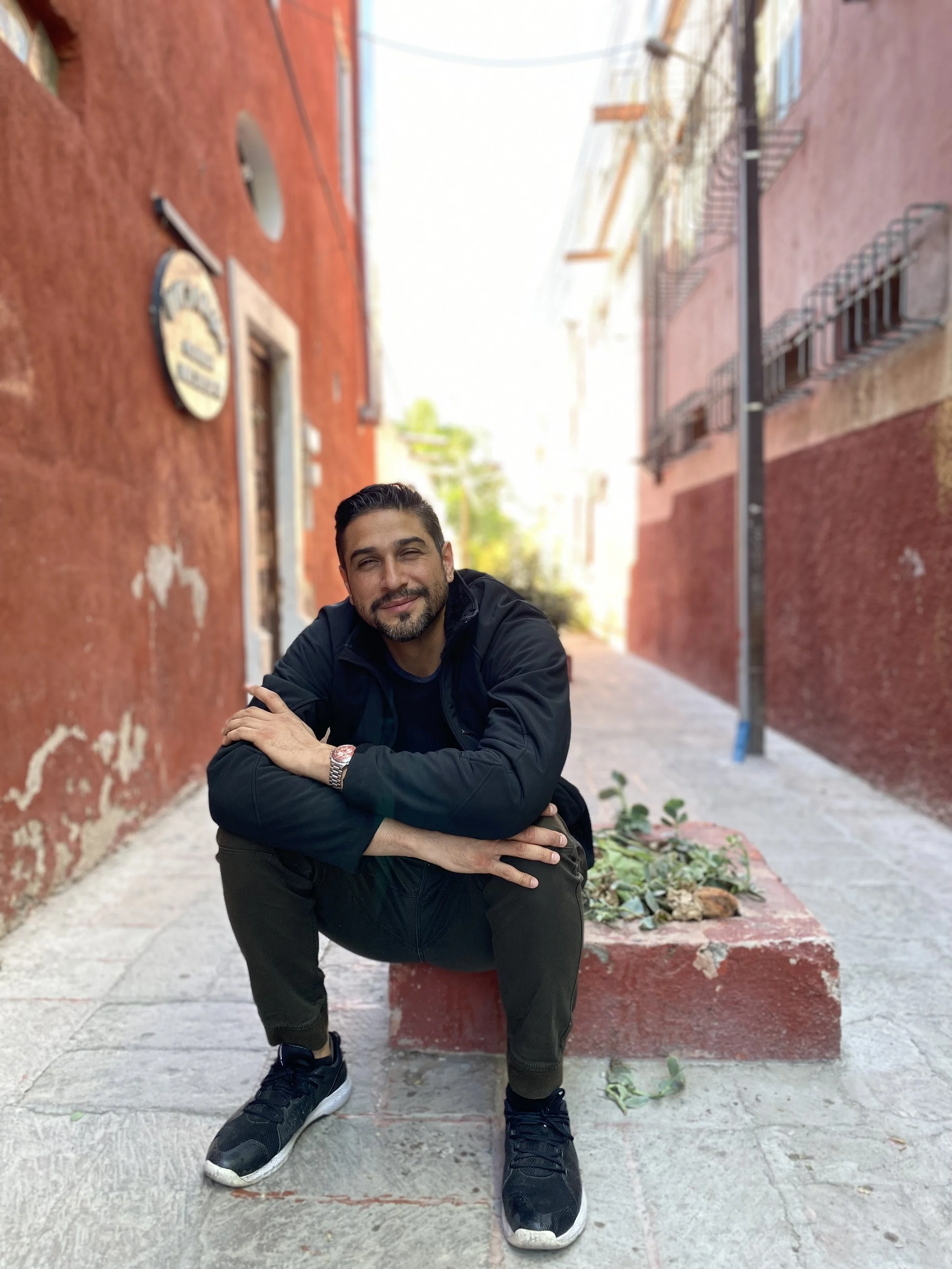 Mauro Falfan sonriente sentado en un banco en una calle estrecha, con paredes de color rojo y edificios a los lados.
