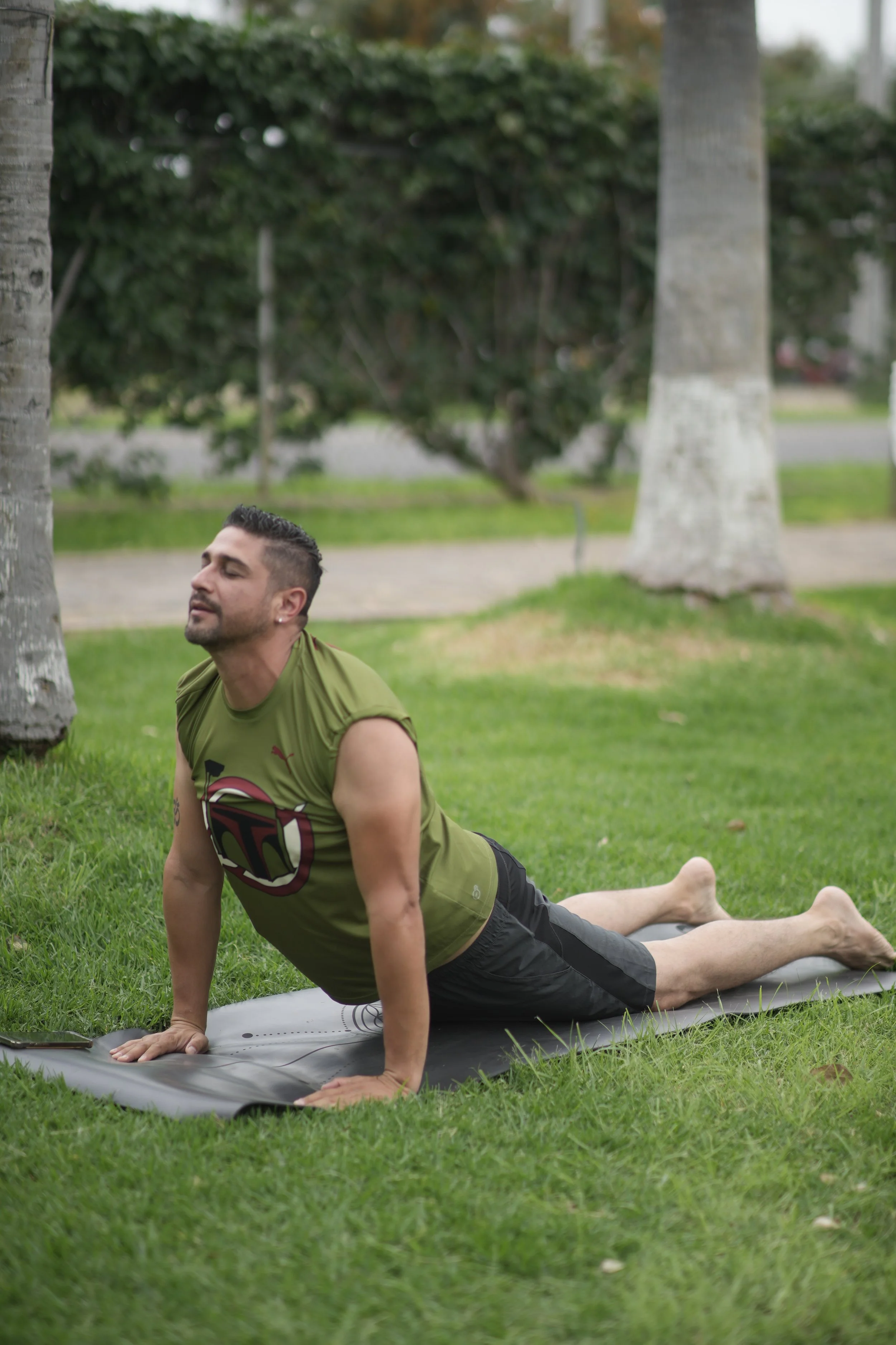 Mauro Falfan haciendo yoga en el parque, en posición de cobra, sobre una colchoneta de ejercicios, rodeado de árboles y césped.