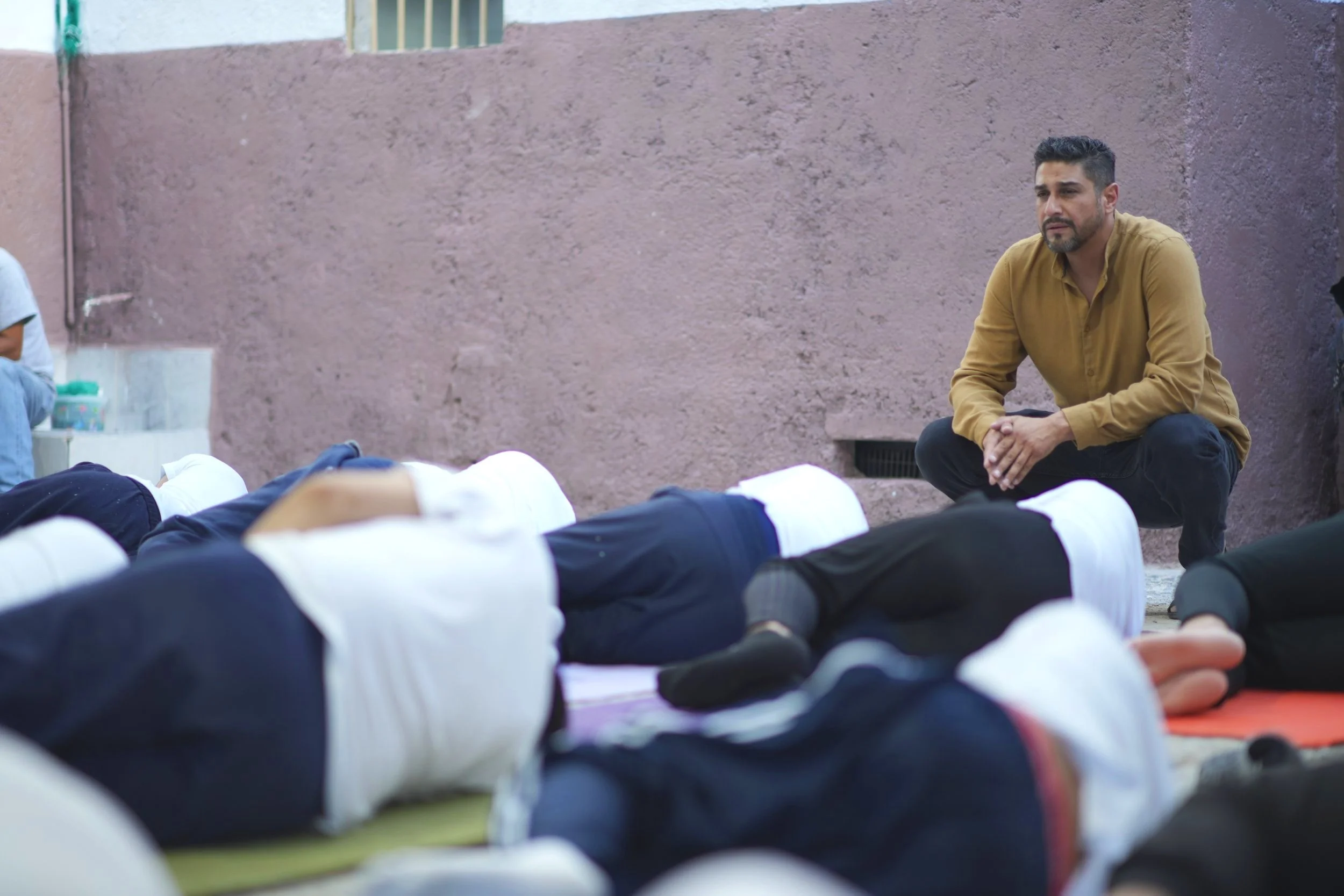 Mauro Falfan obeservando a un grupo de personas que oran en un patio, algunos en posición de oración y cubriéndose el rostro, mientras el hombre los observa de rodillas.