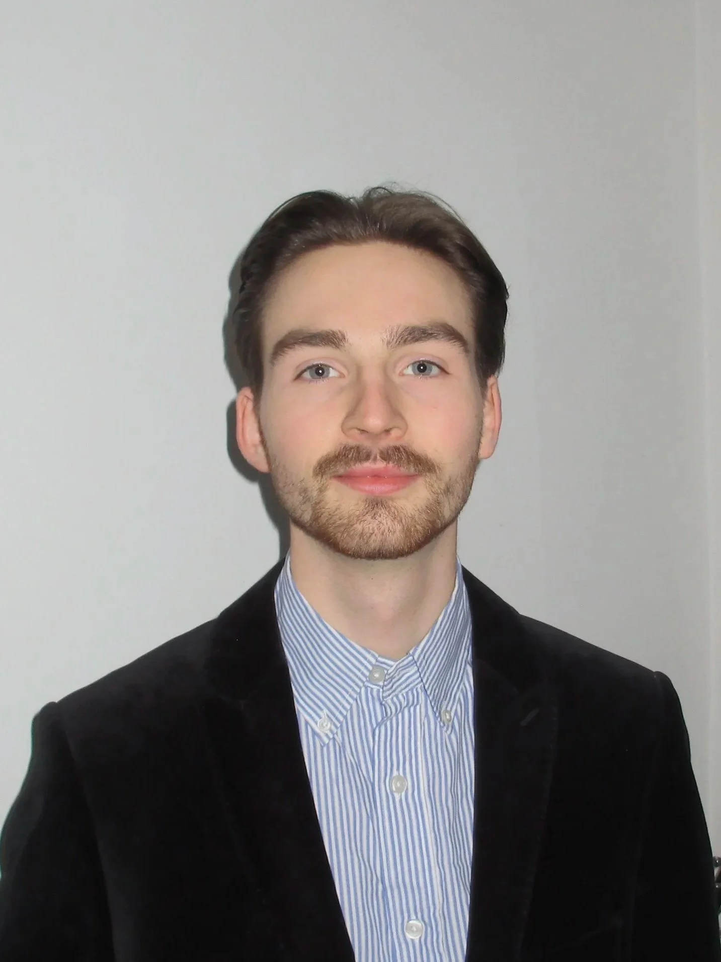 Porträt eines jungen Mannes mit dunklem Haar, Bart und blauen Augen, trägt ein dunkelblaues Jackett und ein hellblau-weiß gestreiftes Hemd, steht vor einem hellgrauen Hintergrund.