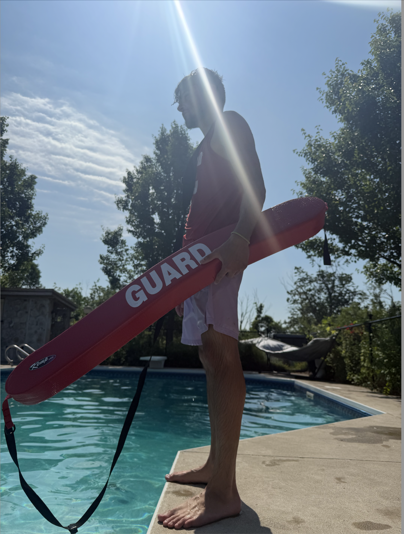 A person standing on the edge of a swimming pool holding a red rescue board labeled 'GUARD,' with trees and a clear blue sky in the background. The sun is shining brightly, creating a lens flare.