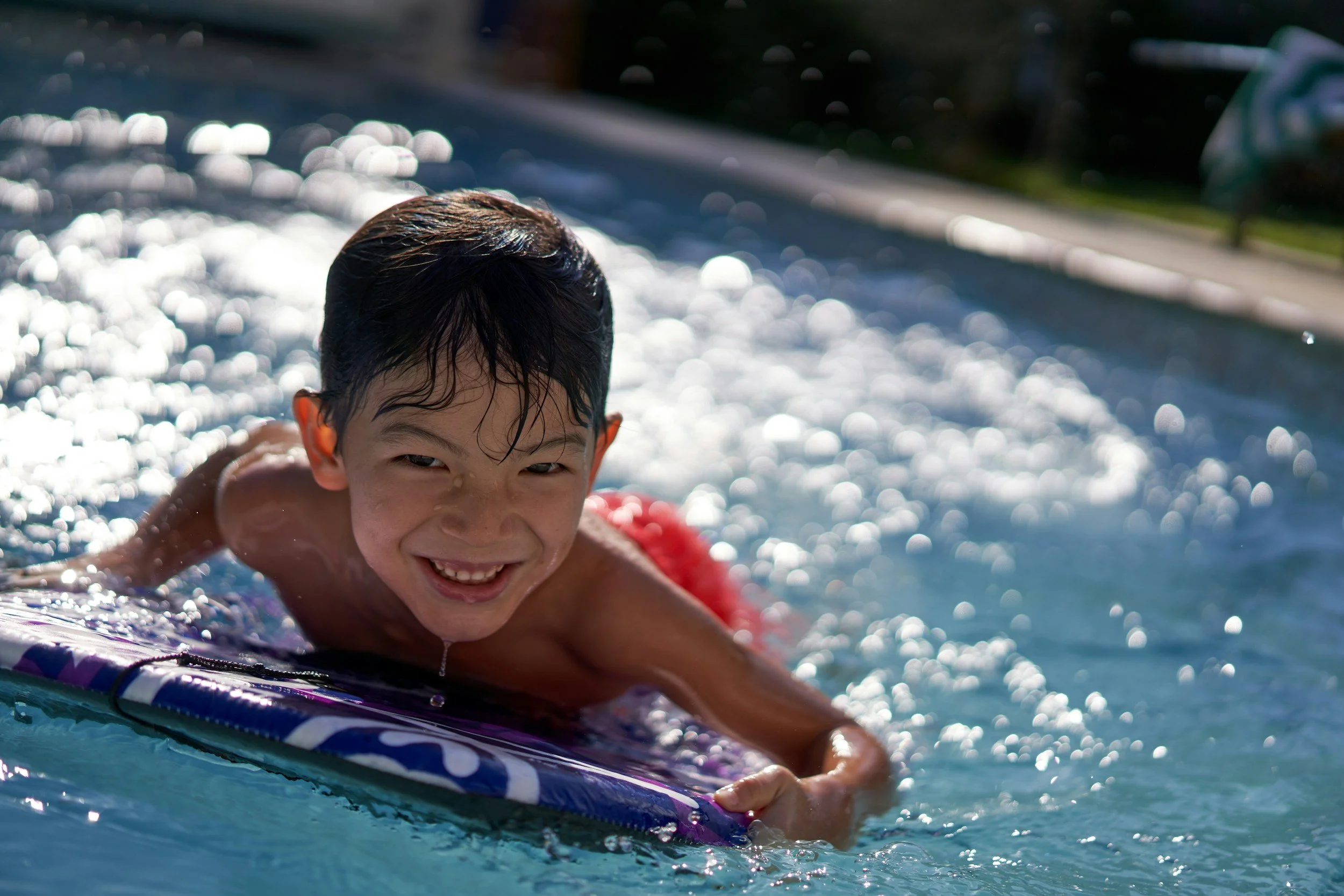 A young boy with wet hair smiling while lying on a surfboard in a swimming pool with sunlight reflecting on the water.