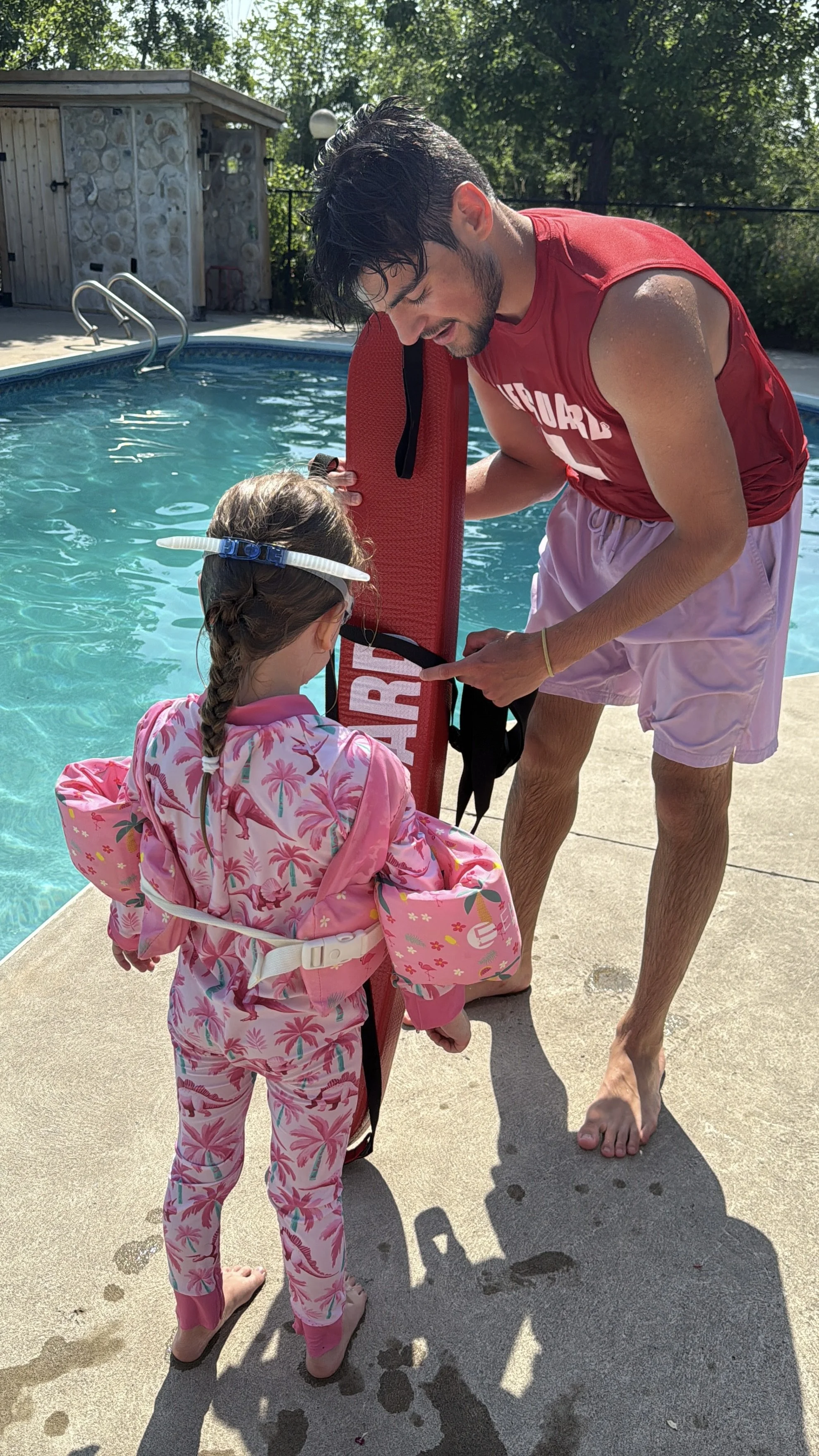 A man and a young girl near a swimming pool, the man helping the girl with a rescue float, both standing on a concrete pool deck with trees in the background on a sunny day.