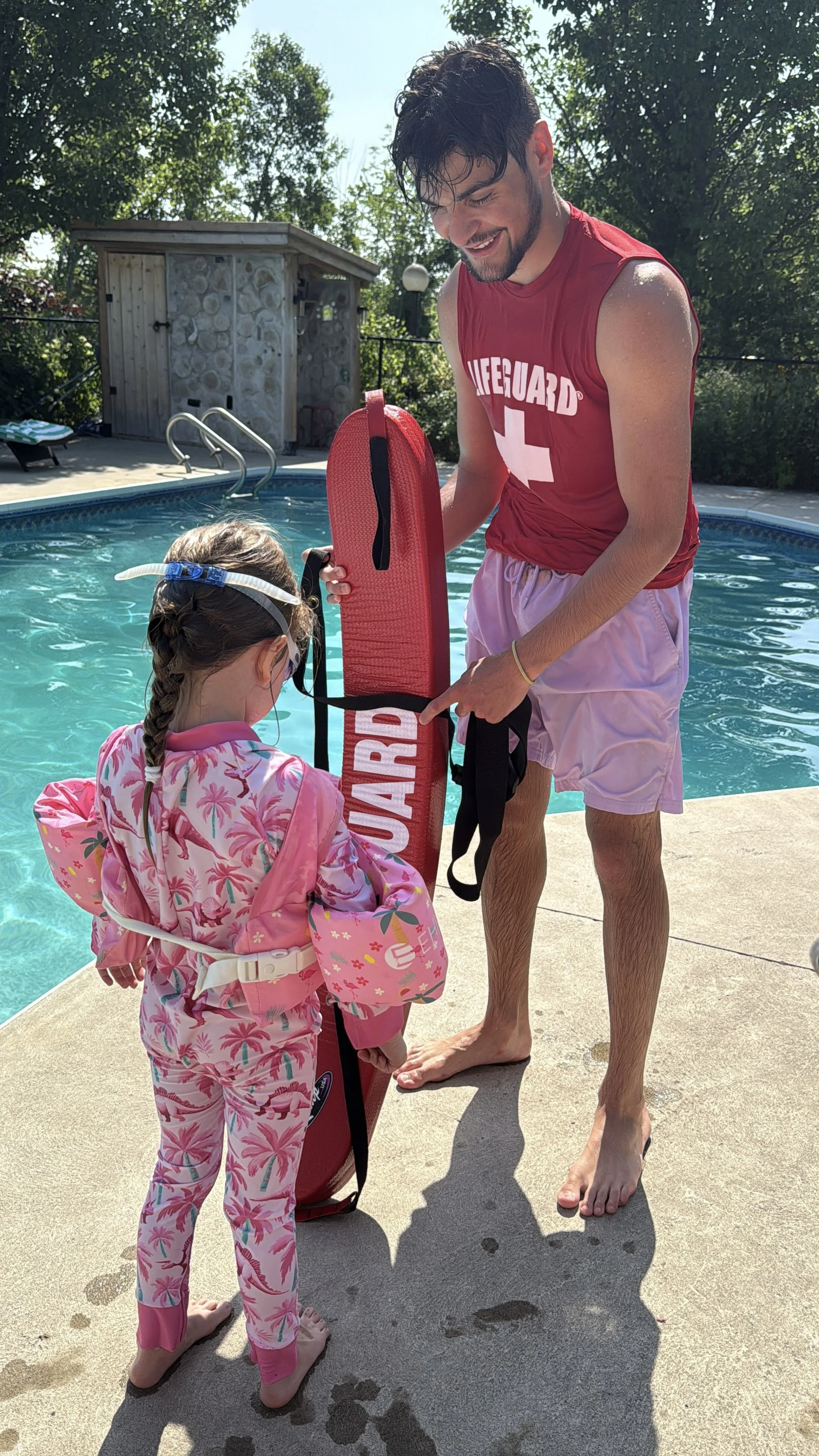 A man and a young girl standing by a swimming pool, preparing for a swimming lesson. The girl is wearing a pink and white floral swimsuit with a life jacket, and the man, dressed in a red Lifeguard shirt and pink shorts, is holding a red rescue backboard.