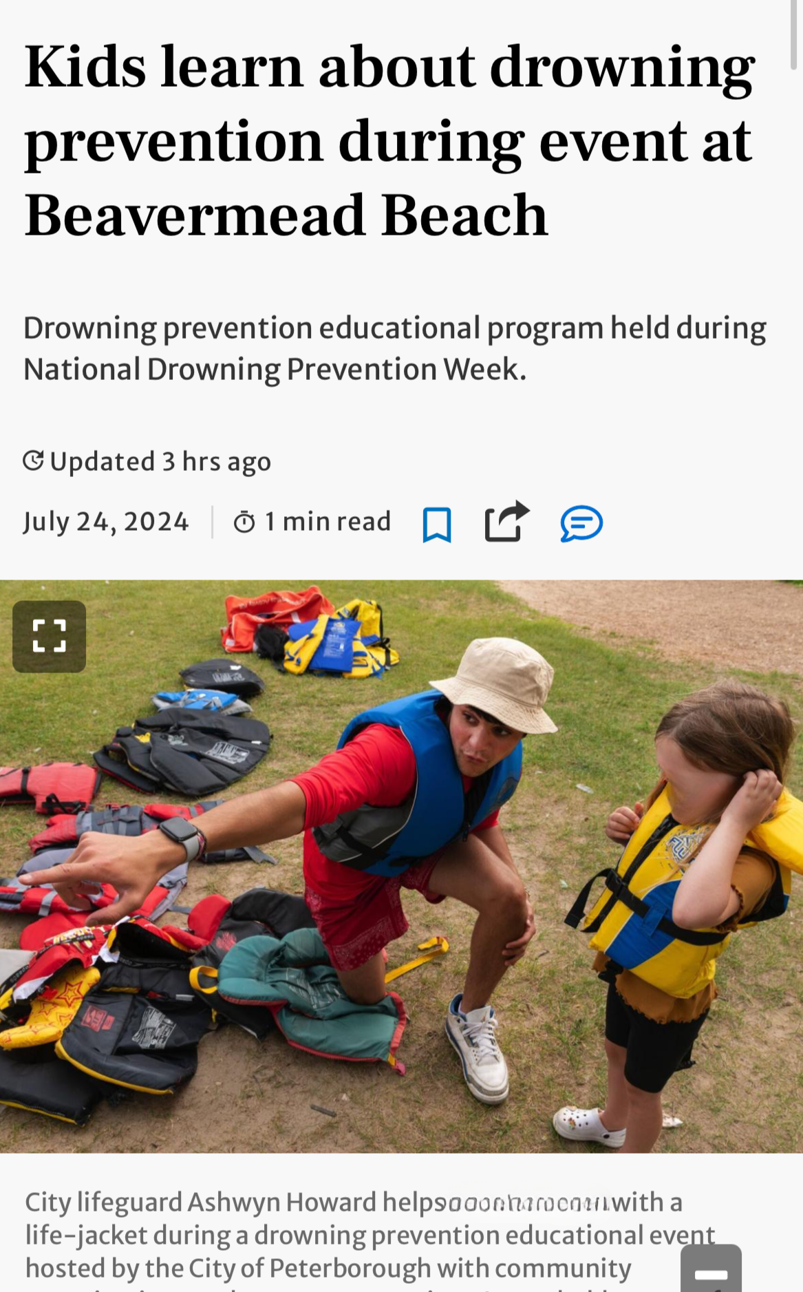 City lifeguard Ashwyn Howard helps a young girl with a life jacket during a drowning prevention educational event at Beavermed Beach, with life jackets laid out on the grass around them.
