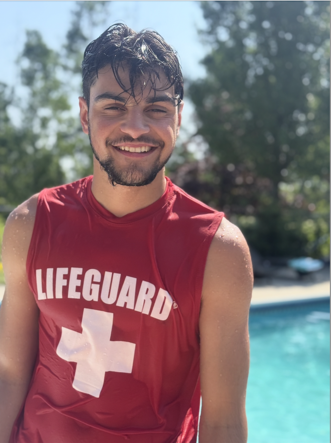 Young man smiling, wet hair, wearing a red lifeguard shirt with a white cross, standing outdoors near a swimming pool with trees in the background.