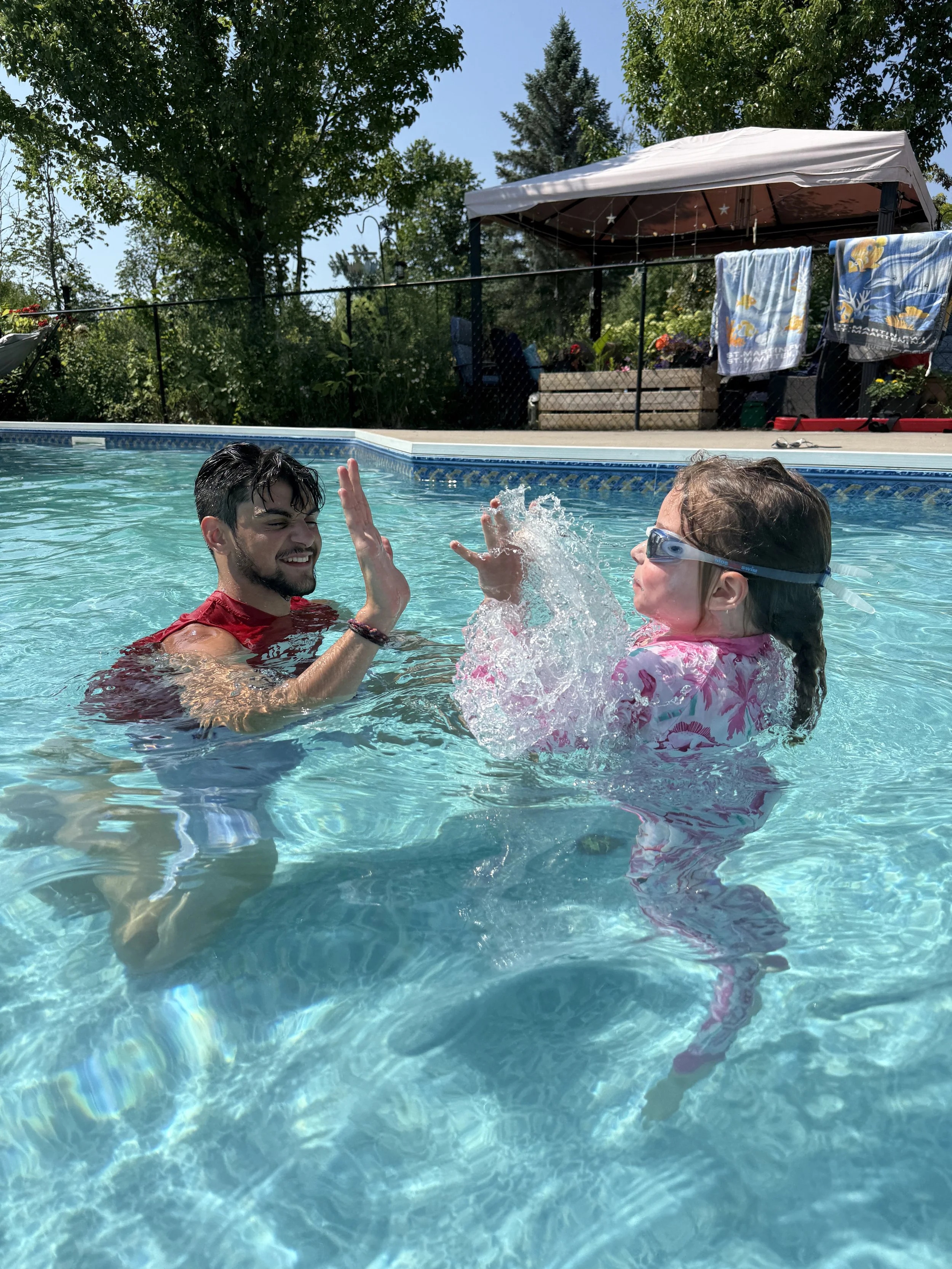 A man and a young girl are in a swimming pool, playing and smiling at each other. The man is giving the girl a high five. The background has trees, a gazebo with towels hanging from a line, and a fence.