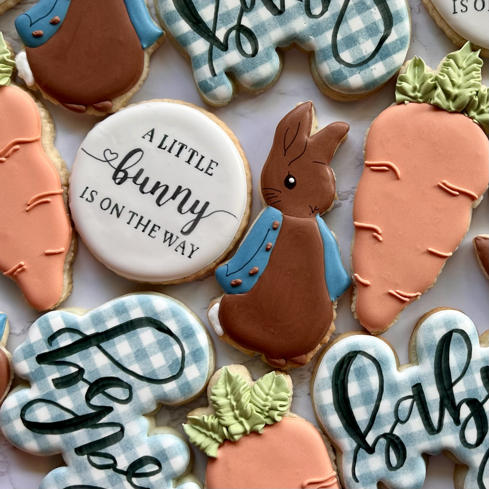 Assorted baby shower cookies, including a Peter Rabbit bunny, carrots, a blue checkered shape with black 'baby' lettering, and a message that reads 'A little bunny is on the way.'