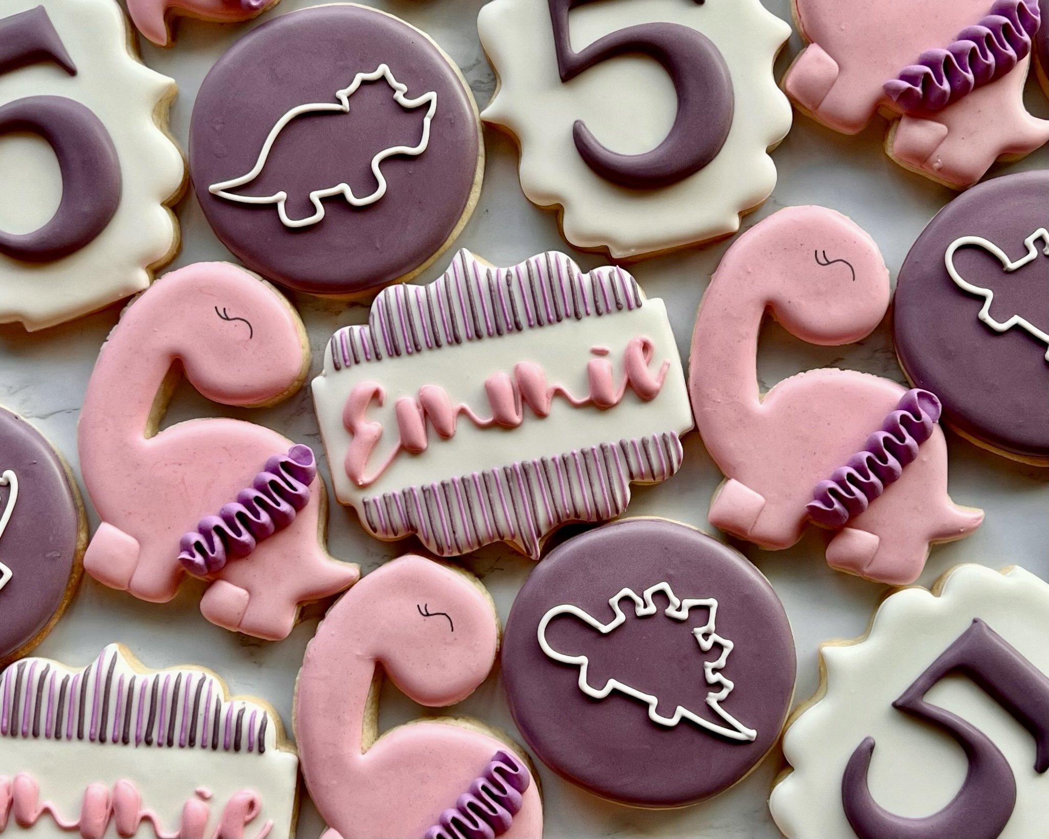 Decorated cookies with dinosaurs, the number 5, and a name plate reading 'Emmie' in pink and purple colors.