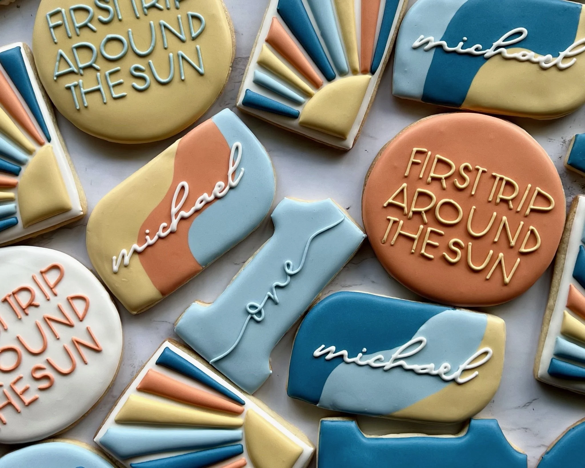Collection of decorated cookies with phrases 'First Trip Around the Sun', name 'Michael', and decorative sun designs in pastel colors.