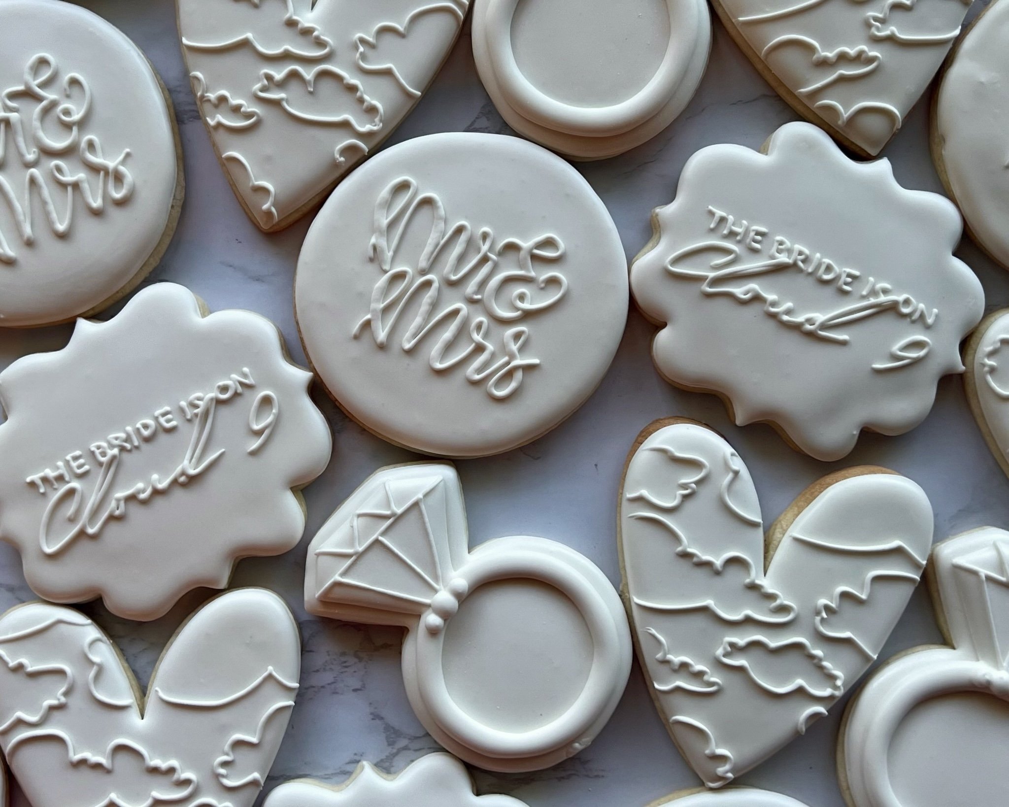 Assorted wedding cookies with white icing, some featuring the words 'Mr. & Mrs.' and 'The bride is on cloud 9' written in cursive, others decorated with clouds, a diamond ring, and a heart with clouds.