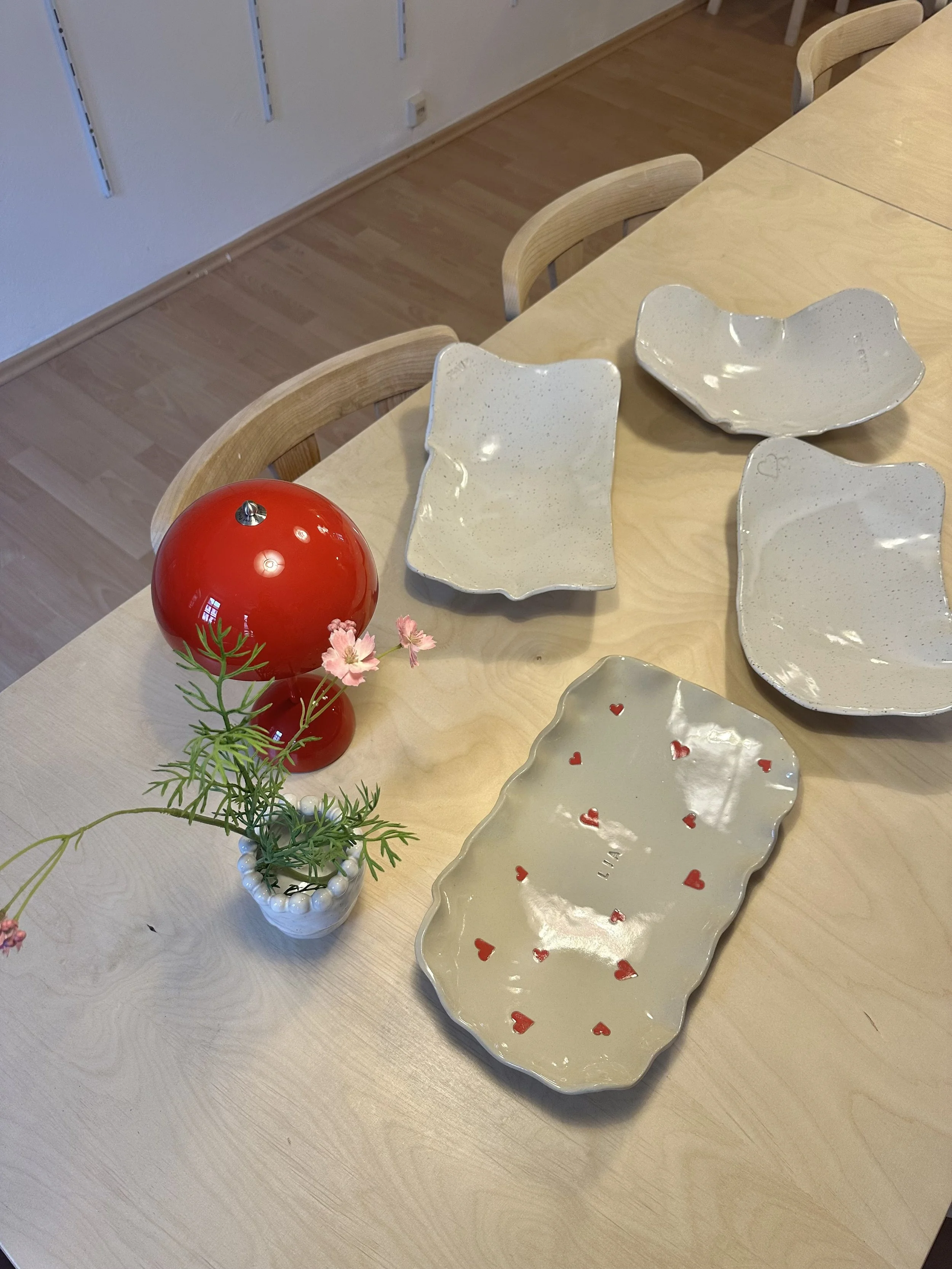Ein Tisch mit weißen Babybauchschalen aus Keramik, einer roten Tischlampe und einem kleinen Blumenstrauß in einer weißen Vase.
