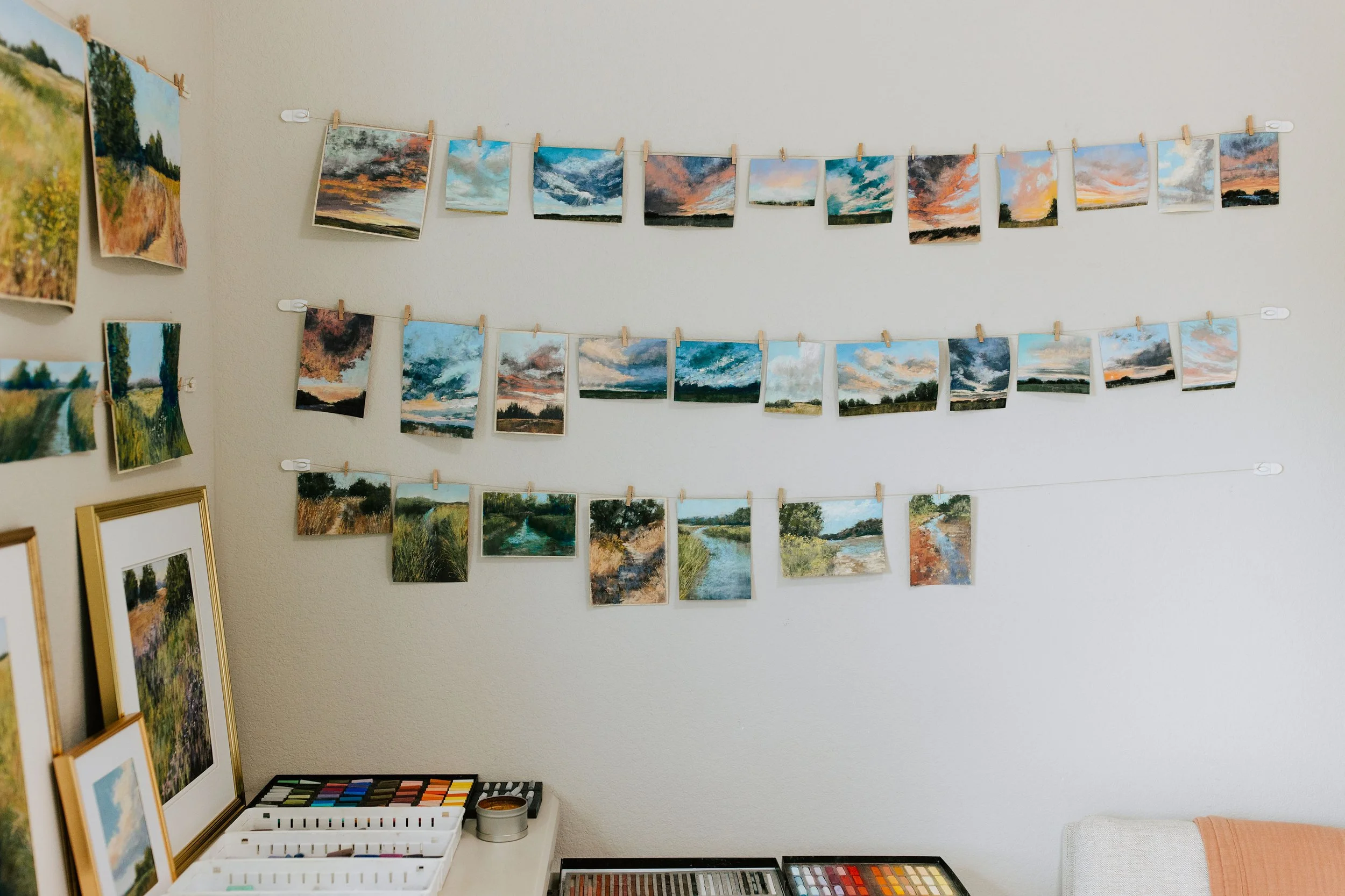 Wall display of small landscape paintings arranged on strings with clothespins, alongside framed artworks and art supplies in a cozy art studio.