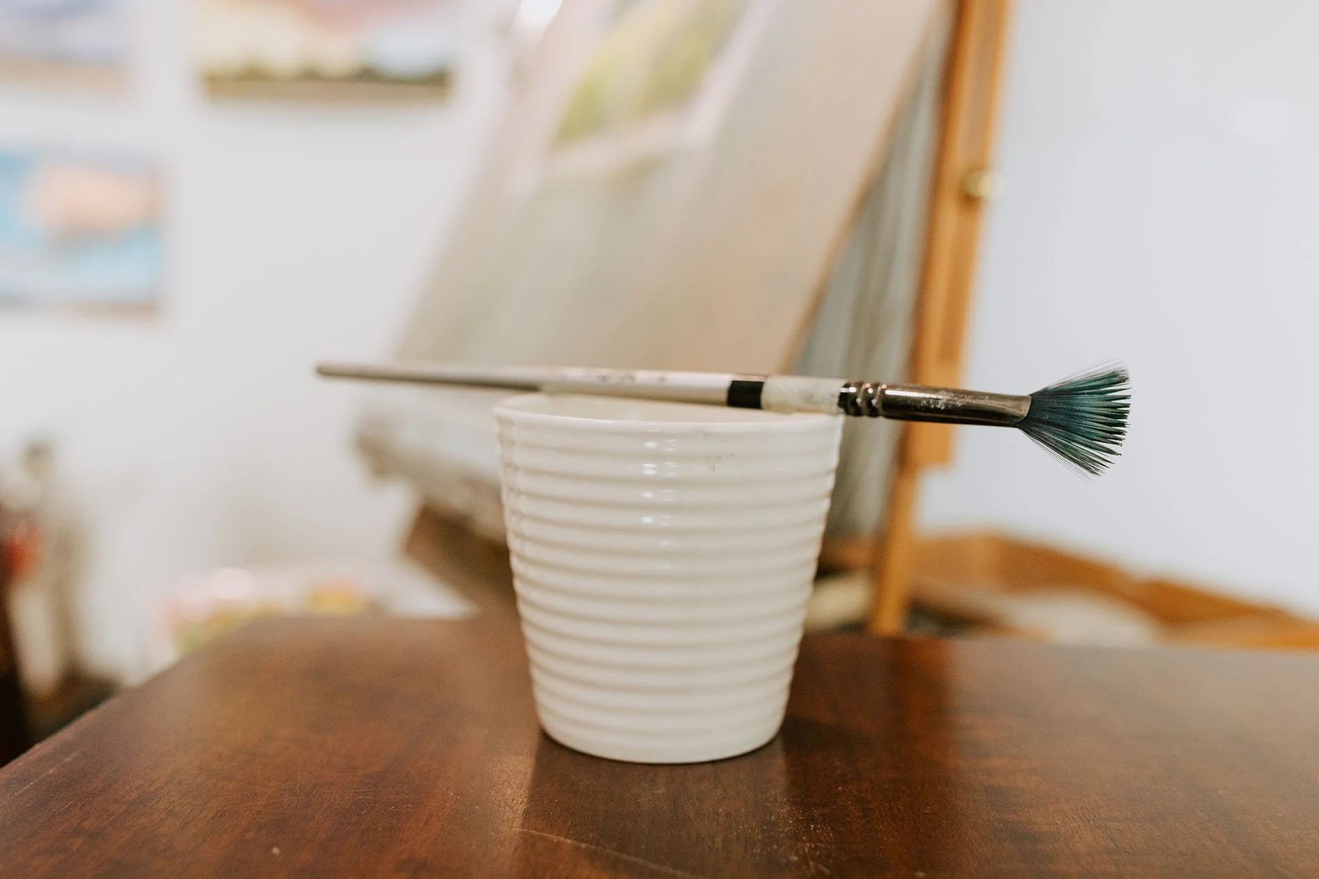 A white ribbed ceramic cup with a paintbrush resting on top, placed on a wooden surface with an easel in the background.
