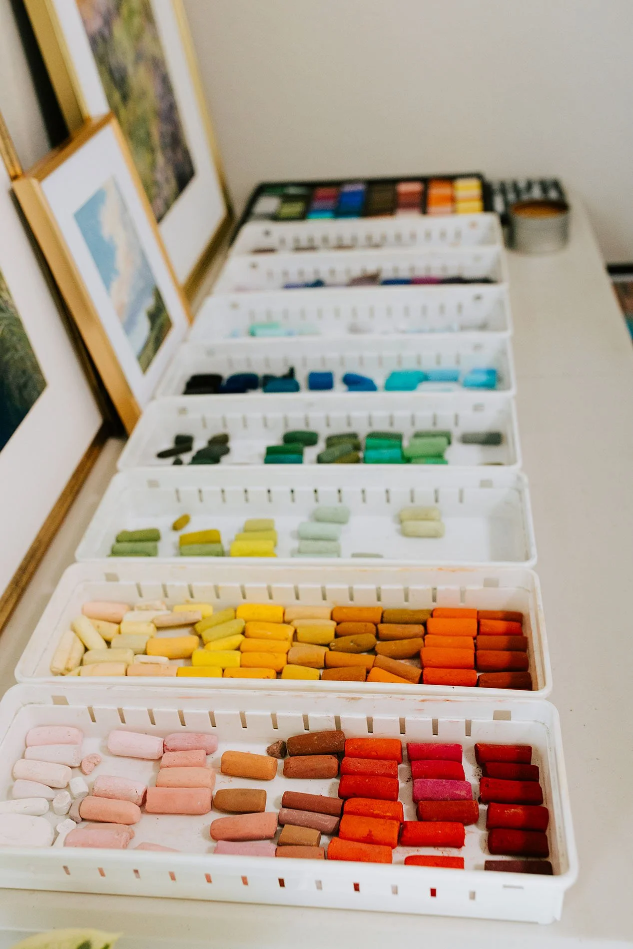 Colorful chalk sticks organized in white trays on a table, with framed landscape photos leaning against the wall in the background.