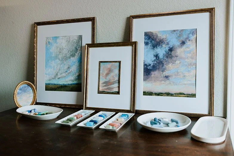 A collection of four framed landscape paintings leaning against a wall on a dark wooden surface, with three white rectangular dishes and a white oval dish filled with soft pastels in front.