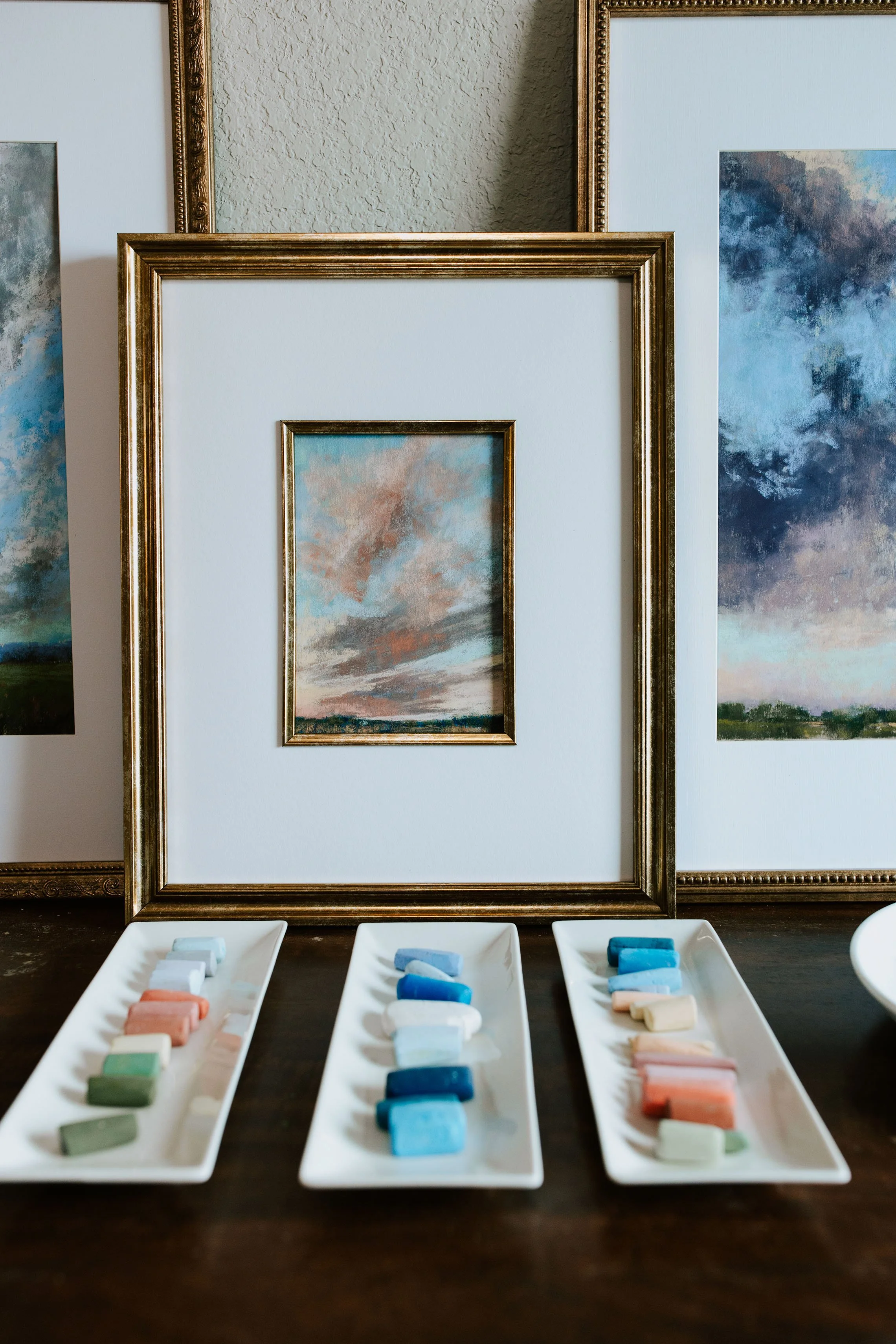 Paintings of sky scenes displayed on a wall, with pastel chalks arranged on white trays in front.