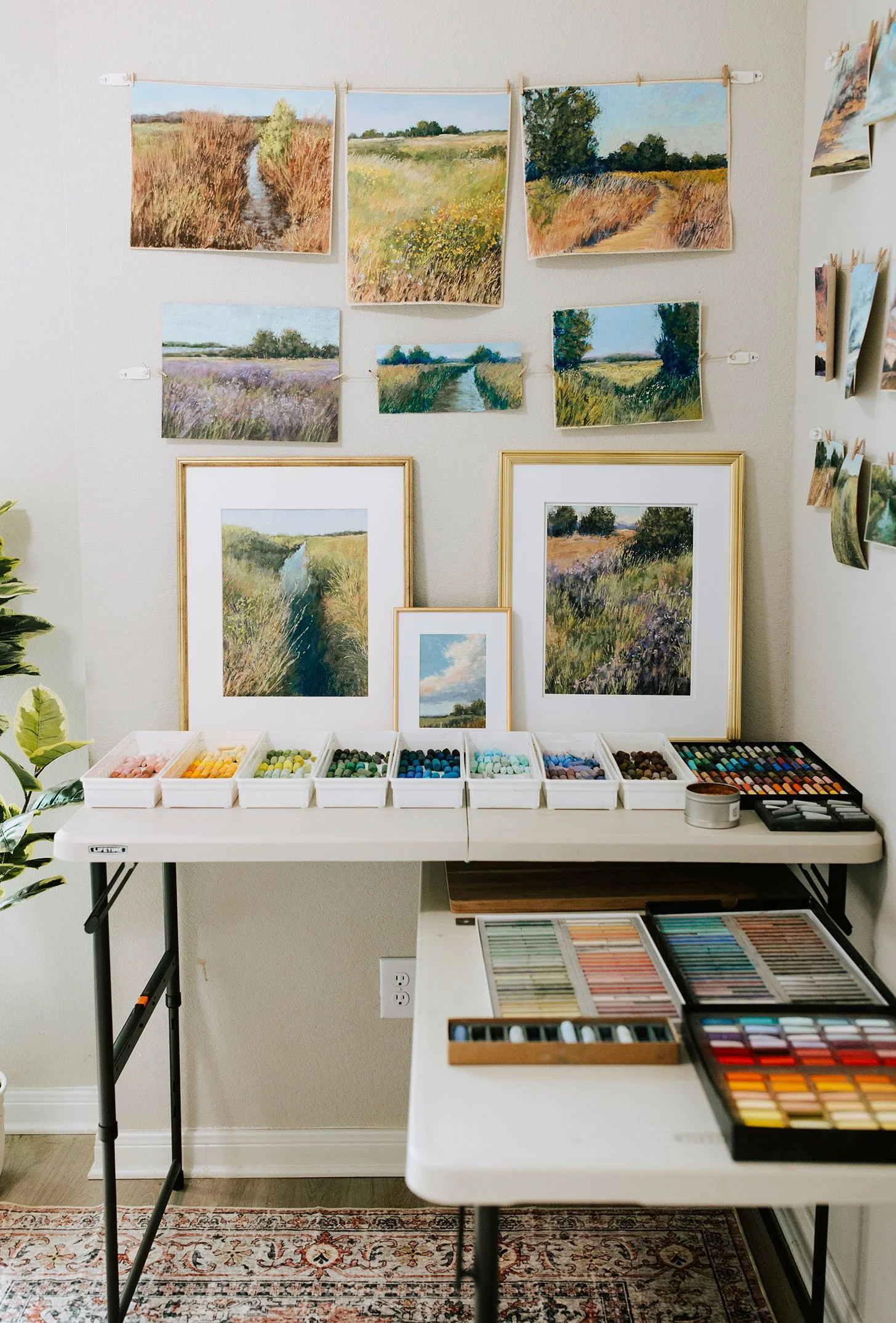 A room displaying multiple framed and unframed landscape paintings of fields and waterways, with colored pastels and art supplies on tables below.