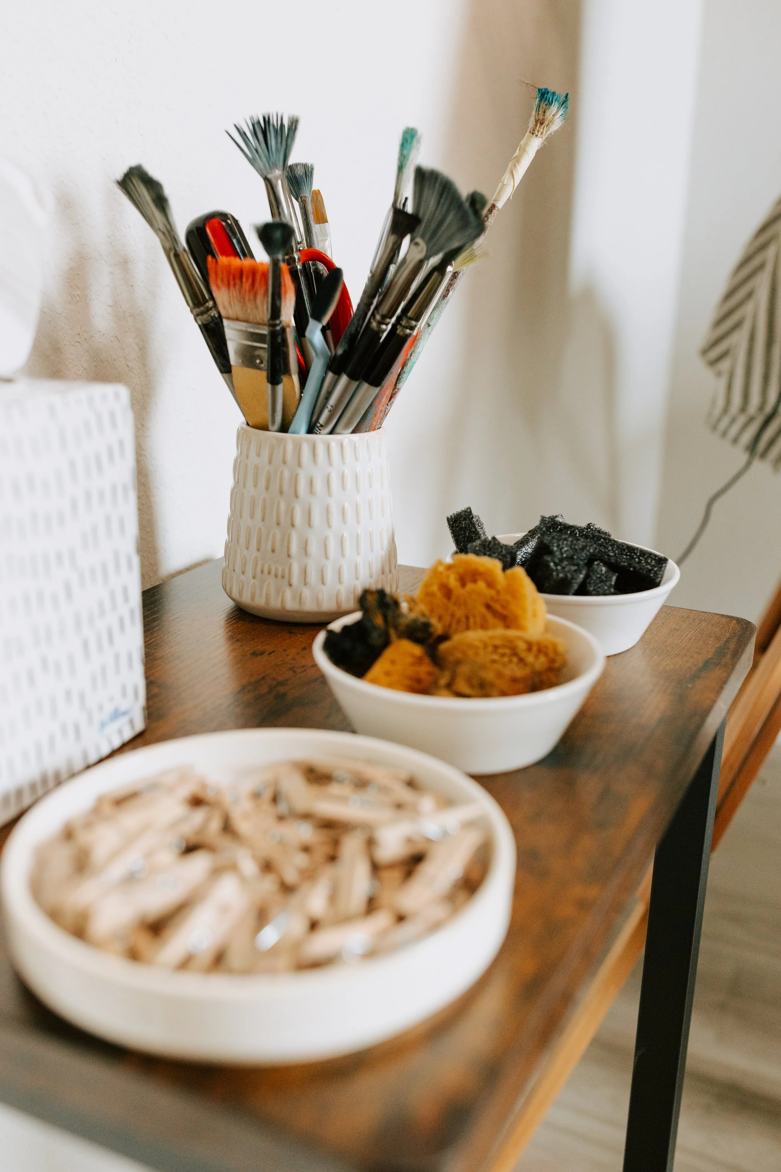 A wooden table with bowls of black and yellow snacks and a white vase holding paintbrushes and art tools.