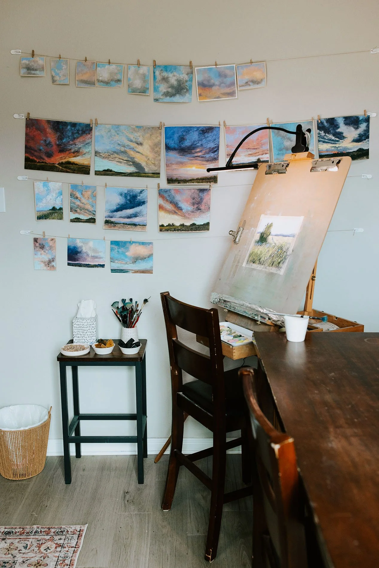 An art studio with a wall displaying multiple small landscape paintings of skies, clouds, and sunsets. A large canvas with an unfinished painting is on an easel, surrounded by art supplies. A small black table holds art tools and snacks, with a woven