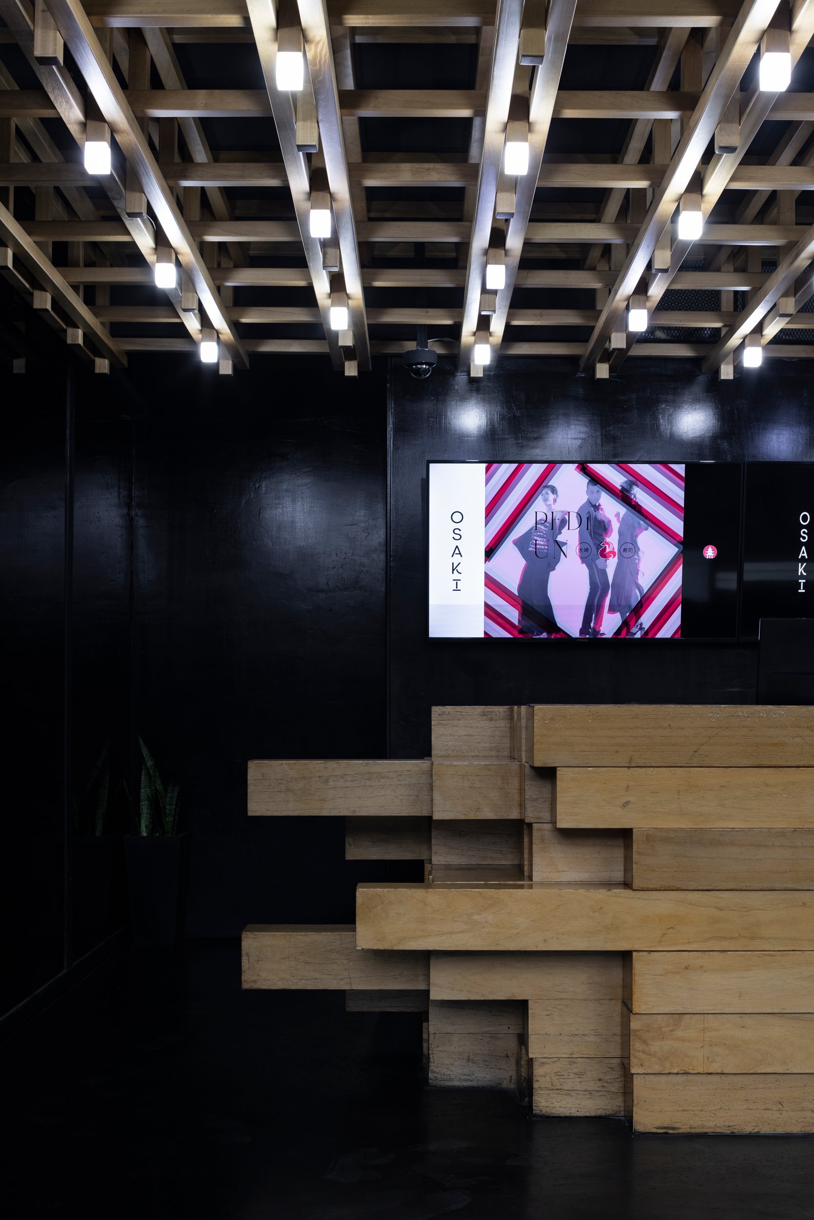 Interior moderno con iluminación empotrada en el techo de madera y una pantalla digital en la pared negra con una imagen de tres personas y el texto 'OSAKI'.