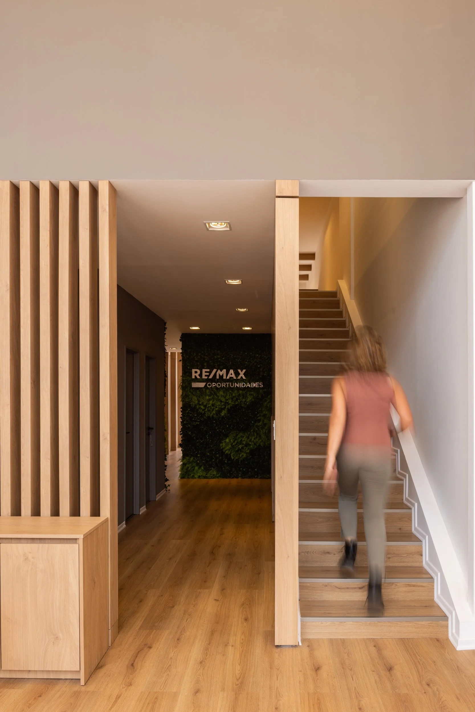 Interior de oficina con pared de plantas, escaleras de madera y señal de RE/MAX OPOTUNIDADES en el fondo