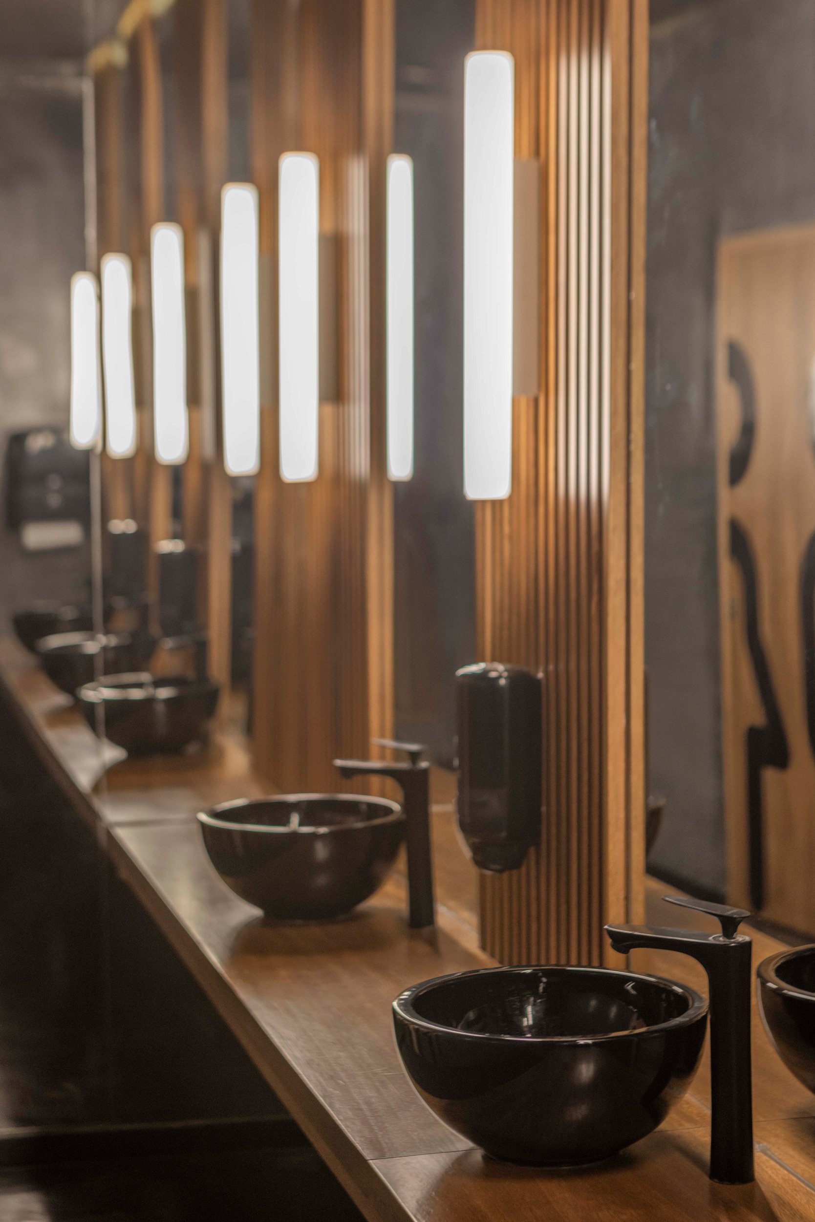 Lavabos y espejos en un baño público moderno con iluminación empotrada y decoraciones en madera.