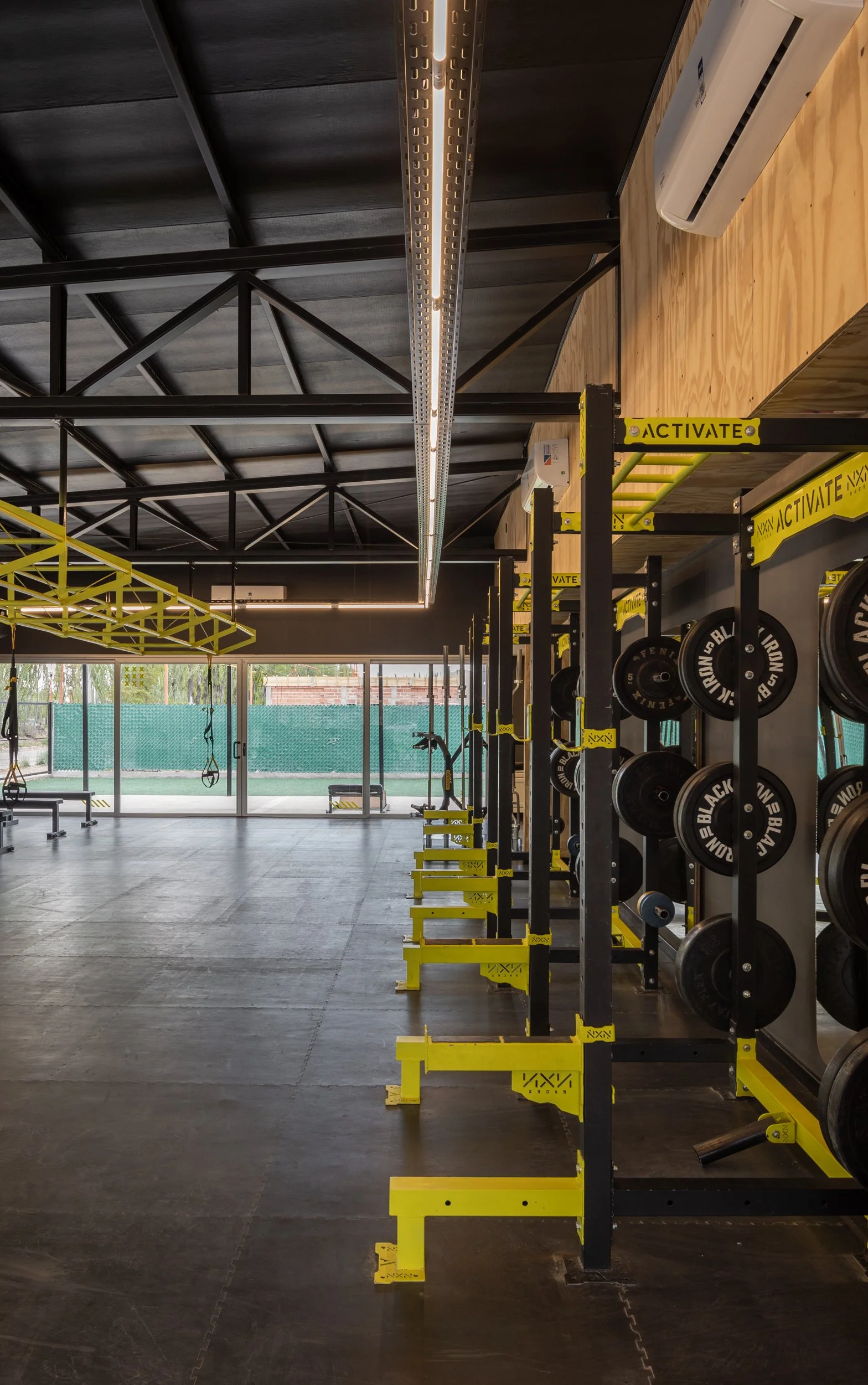 Interior de gimnasio con pesas, barras y equipos de ejercicio, ventanas y vista al exterior.