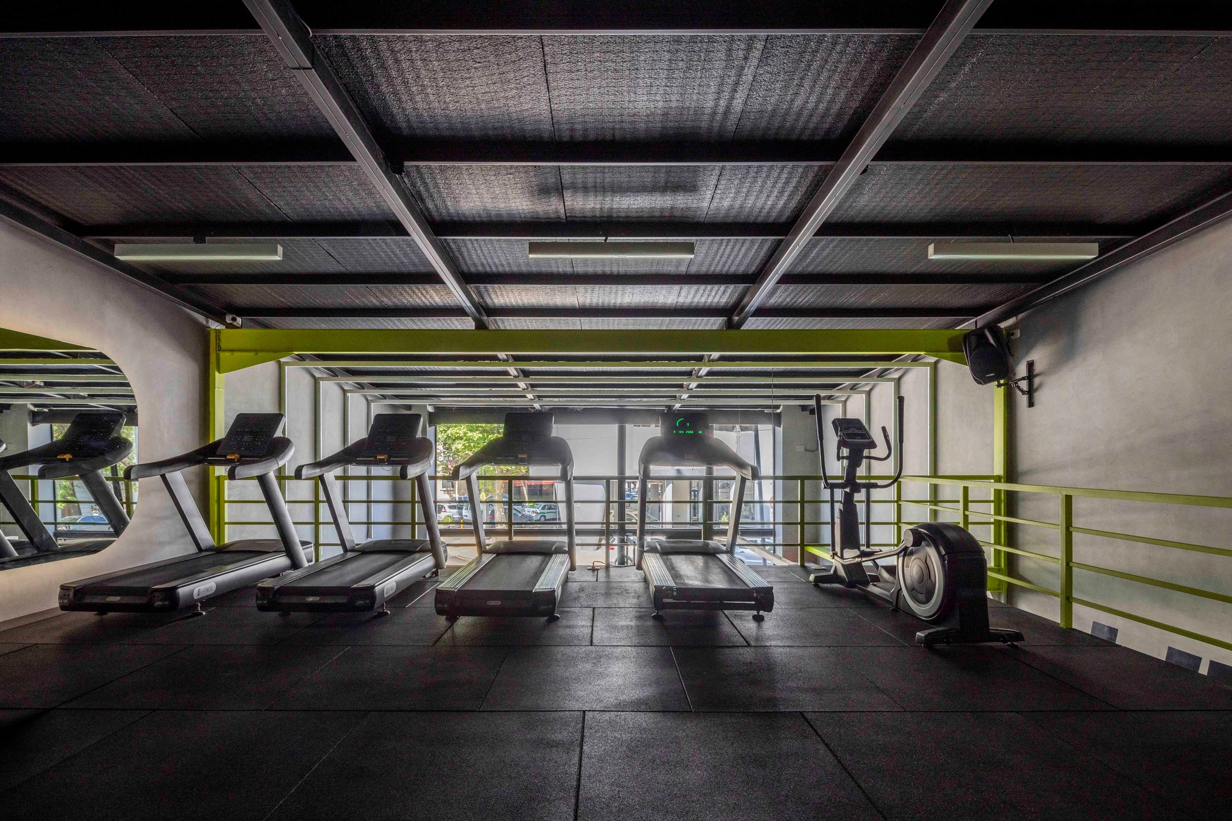 Sala de gimnasio con varias cintas de correr, máquina de elíptica y espejos en la pared, con luz natural entrando por grandes ventanas en el fondo.