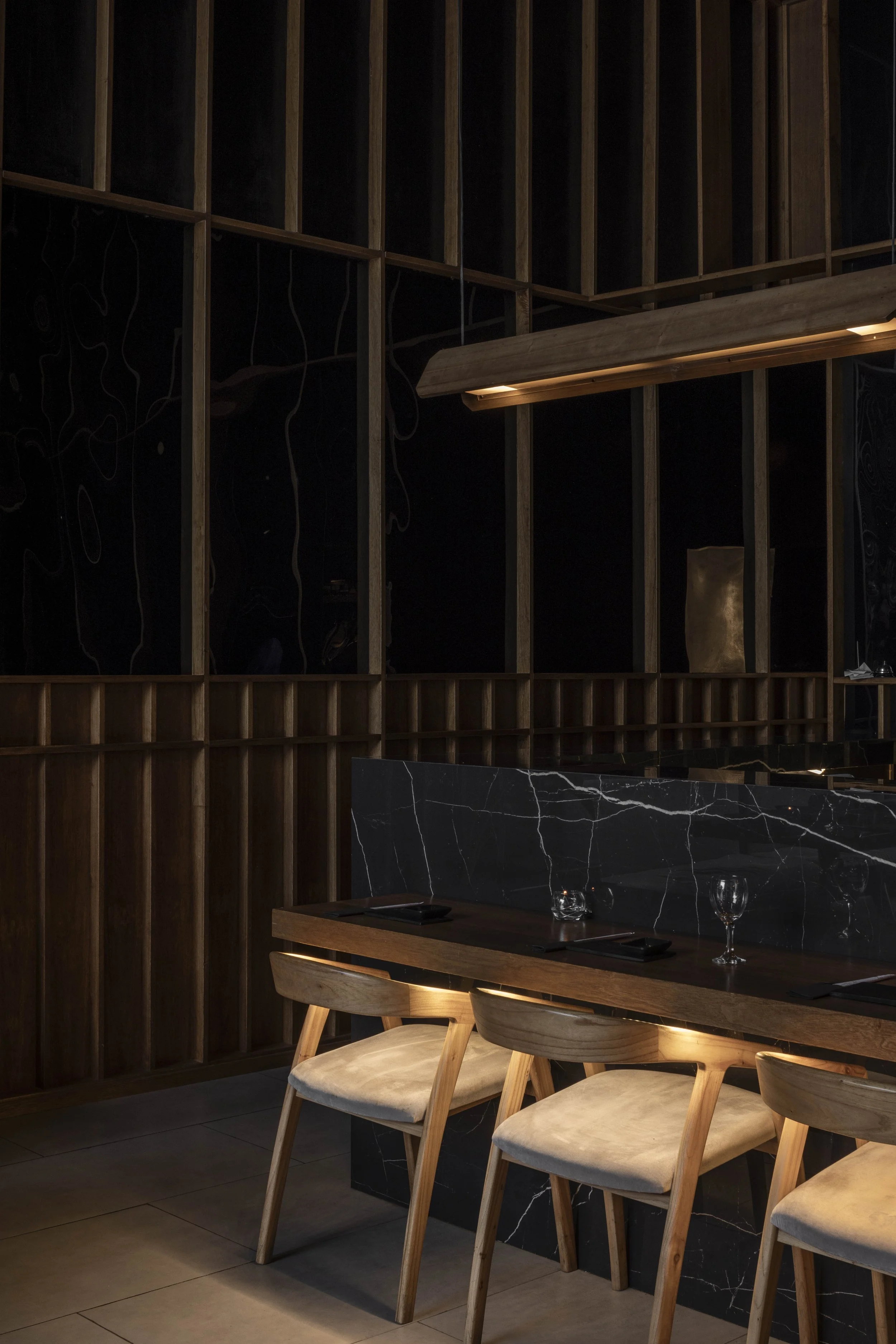 Interior de un restaurante con mesas de madera, sillas de madera con cojines claros, pared de madera oscura, divisiones de mármol negro con vetas blancas y lámpara suspendida de madera.