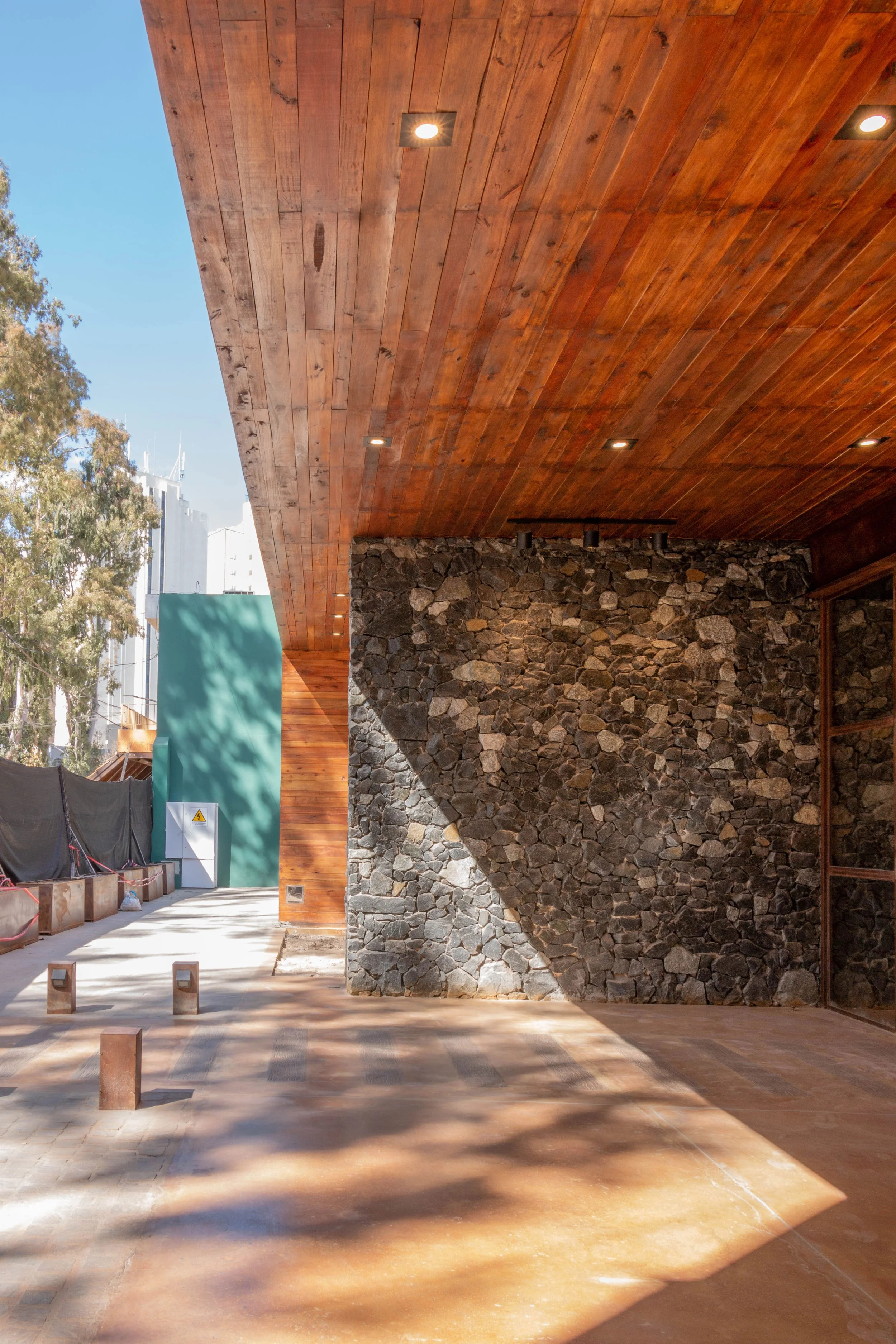 Espacio exterior con techo de madera, pared de piedra y piso de ladrillos, con sombra y árboles en el fondo.