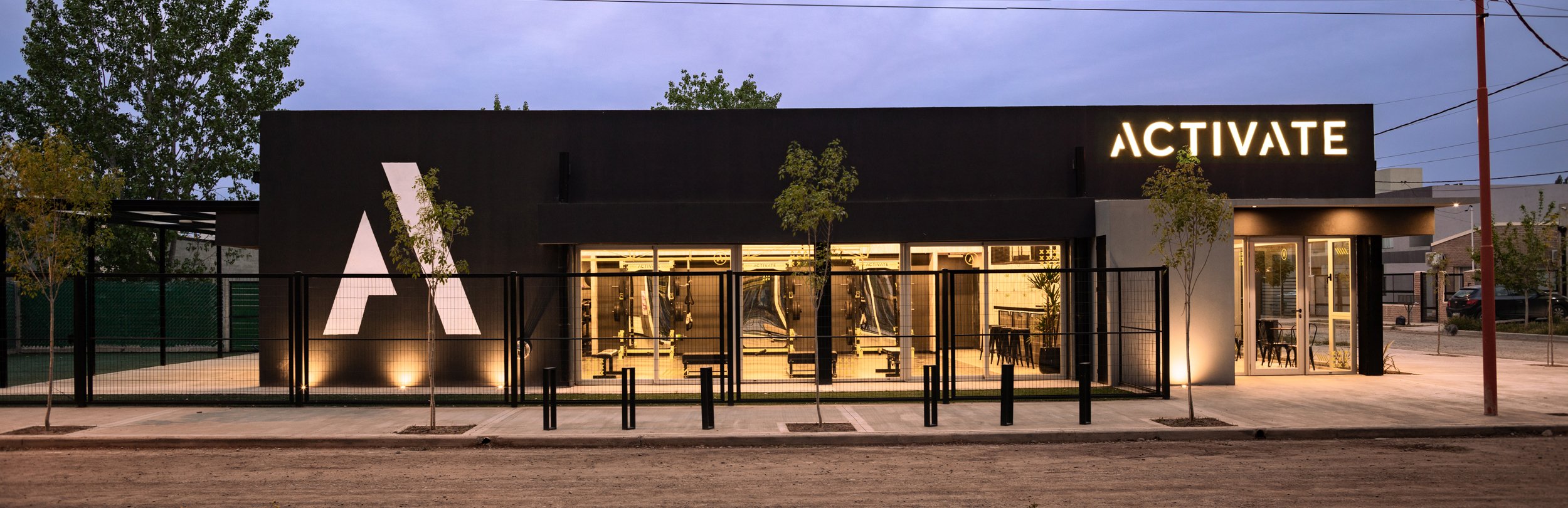 Fachada de un gimnasio moderno llamado ACTIVATE con luces en la noche, con ventanas grandes y área exterior con árboles y vallas negras.