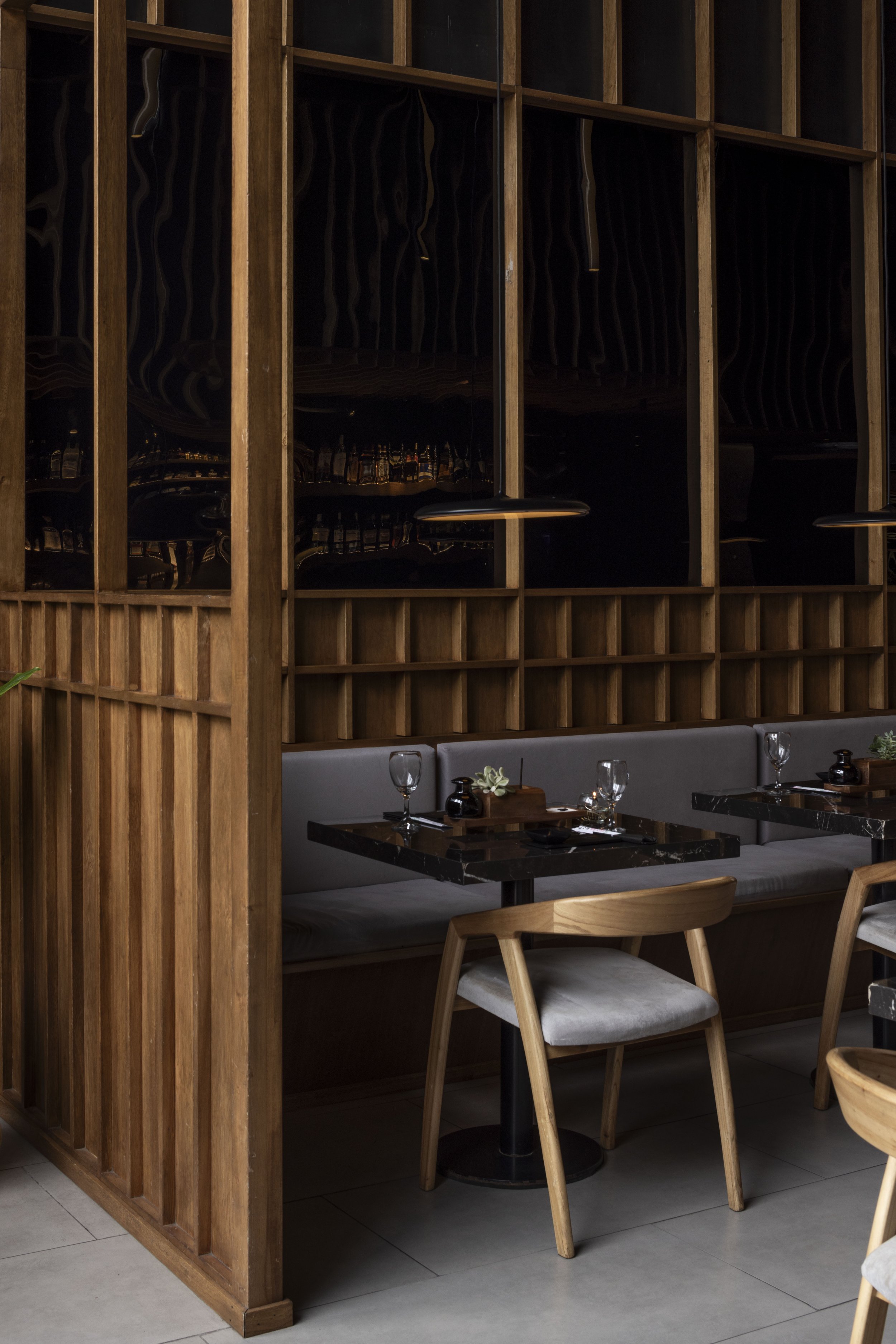 Mesa en un restaurante moderno con copas de vino, servilletas y pequeños jarrones con plantas, además de sillas de madera y un fondo decorativo de madera y paneles oscuros.