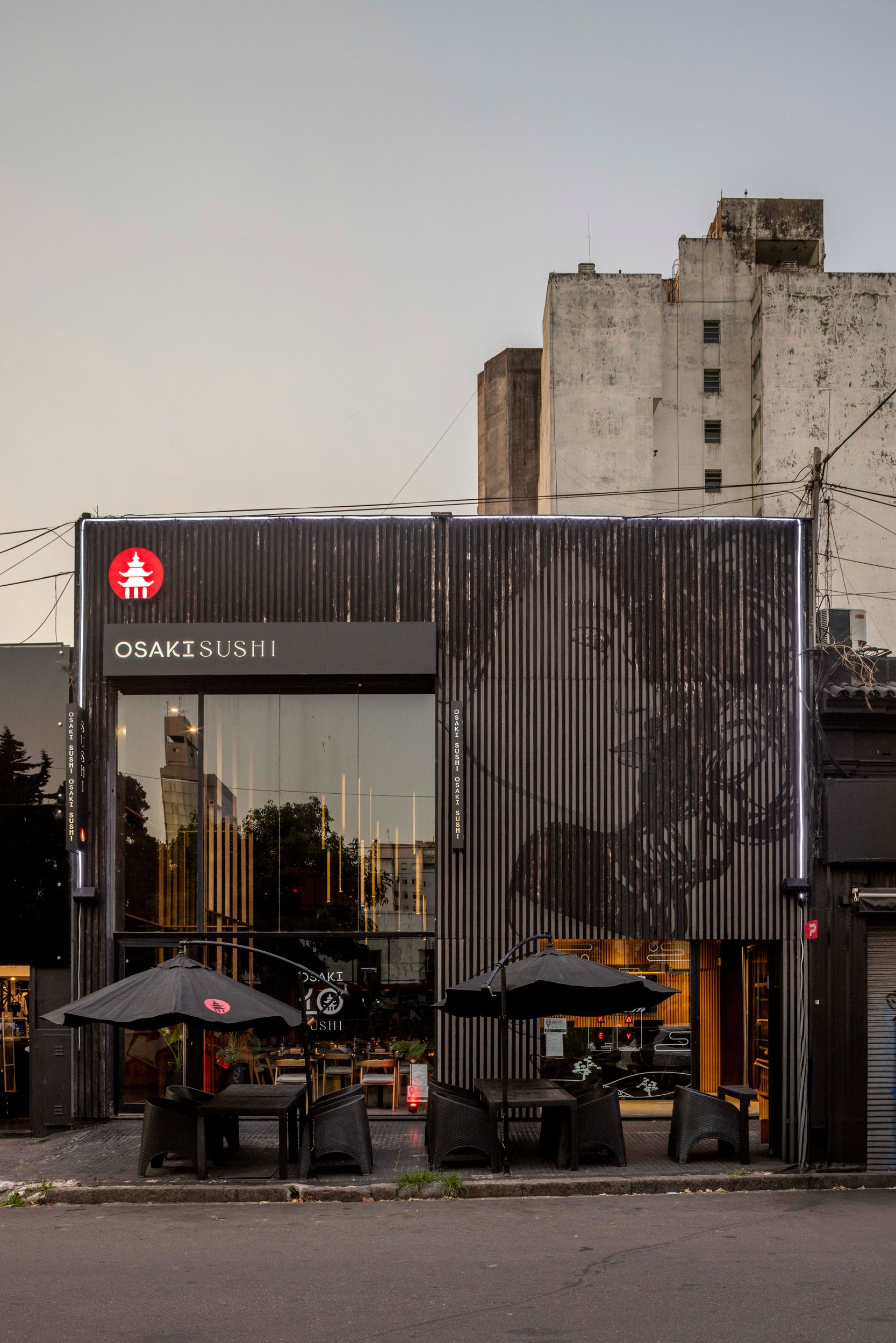 Exterior de un restaurante llamado Osaka Sushi con mesas y sillas en la acera, toldos y un gran cartel con un logotipo rojo y negro.