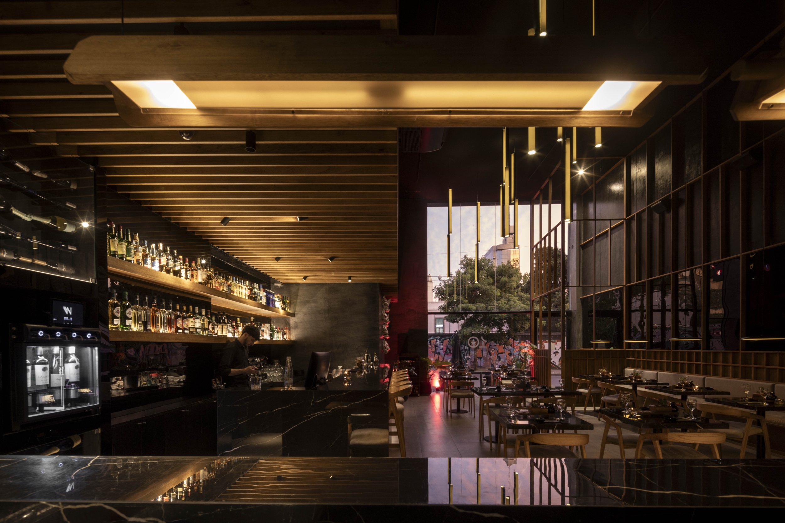 Interior de un restaurante elegante con barra de bebidas, mesas con cubiertos y una gran ventana que muestra árboles y edificios en el exterior.
