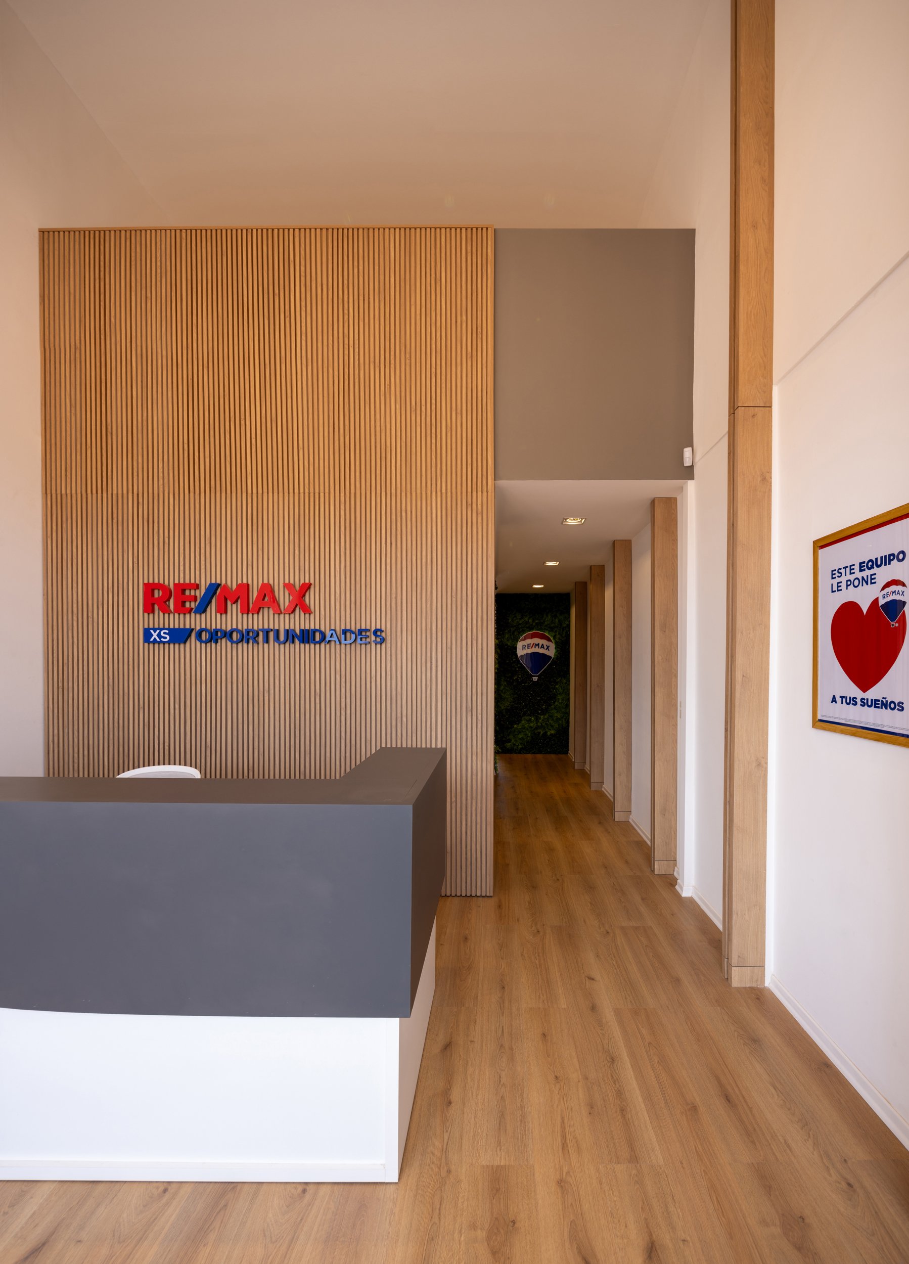 Recepción de oficina con señal de RE/MAX y anuncio de oportunidades, interior con suelo de madera, paredes blancas, decoración moderna y puertas de madera.