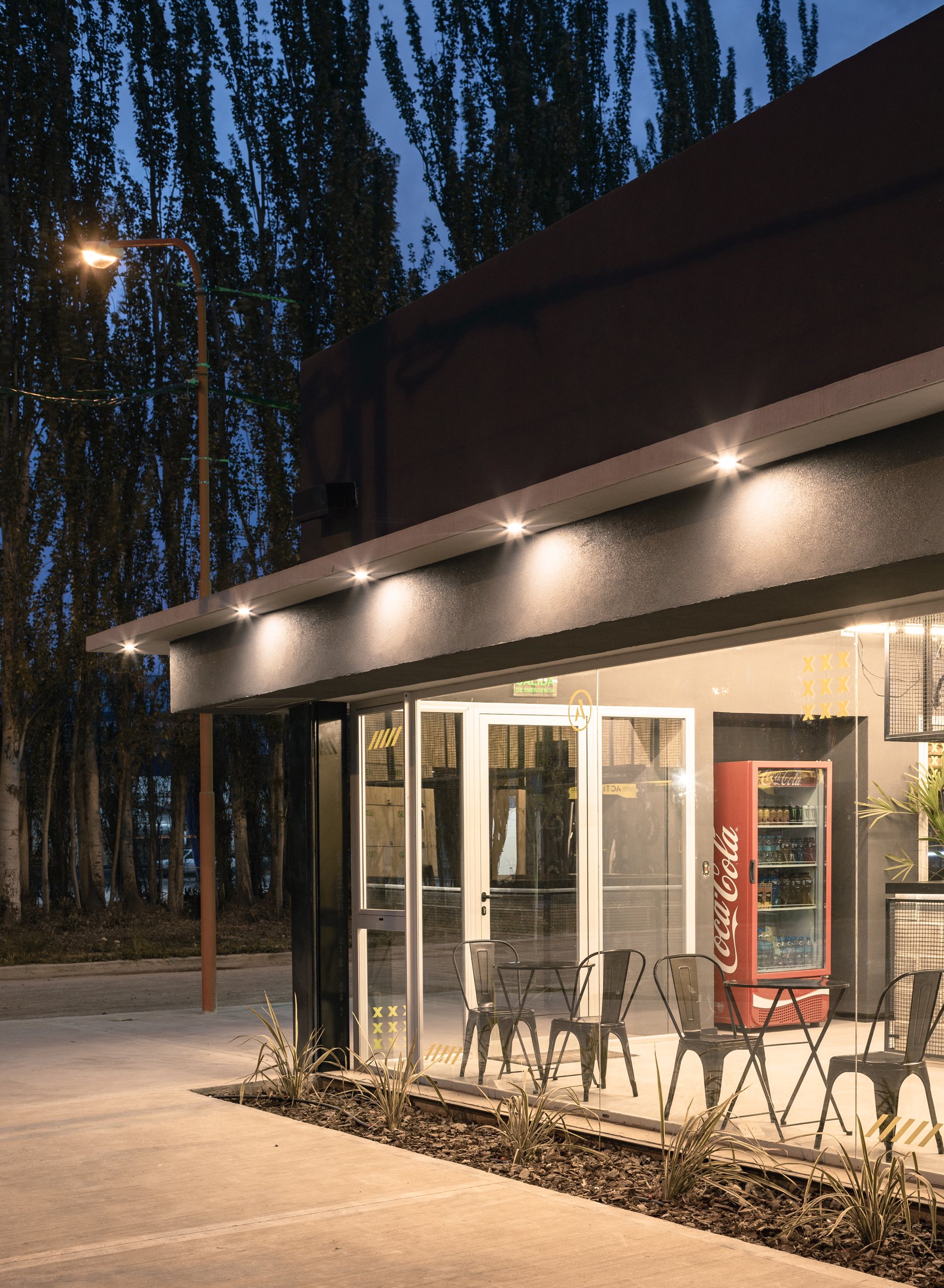 Una tienda de alimentos y bebidas con vallas de metal y refrigerador de Coca-Cola frente a la entrada, en una noche con iluminación artificial