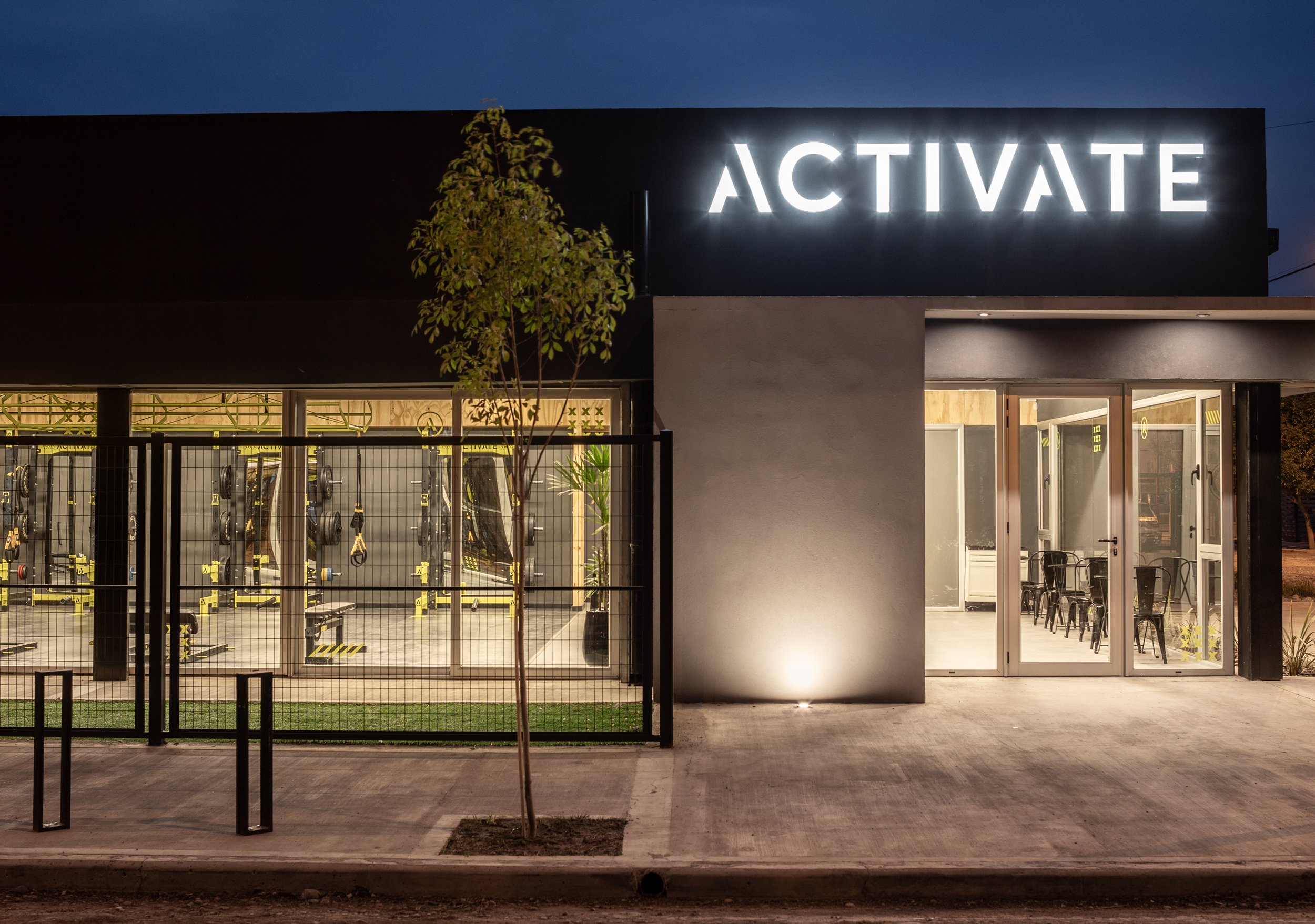 Edificio moderno con cartel de neón que dice 'ACTÍVATE', vista de gimnasio con pesas y equipo de ejercicio visible a través de ventanas de vidrio, a la luz de la noche.