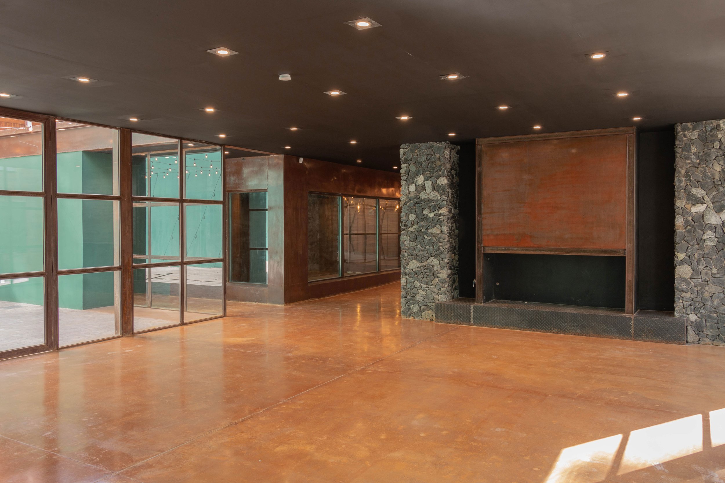 Interiores de un salón amplio con paredes de piedra, suelo de concreto pintado en color marrón y paredes de vidrio con marcos de madera. La decoración es moderna con iluminación empotrada en el techo.
