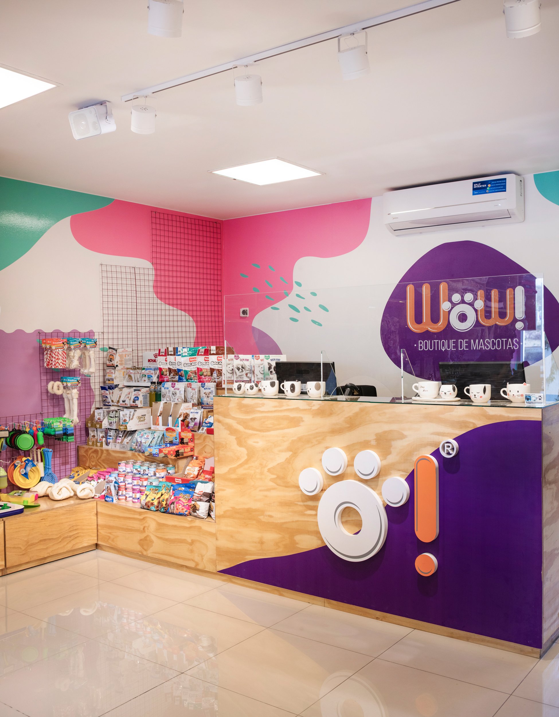 Mostrador en tienda de mascotas con un cartel que dice 'WOW! Boutique de Mascotas' y exhibe tazas con logos, con juguetes y productos para mascotas en estantes de madera, paredes decoradas con manchas de colores y rejillas metálicas.