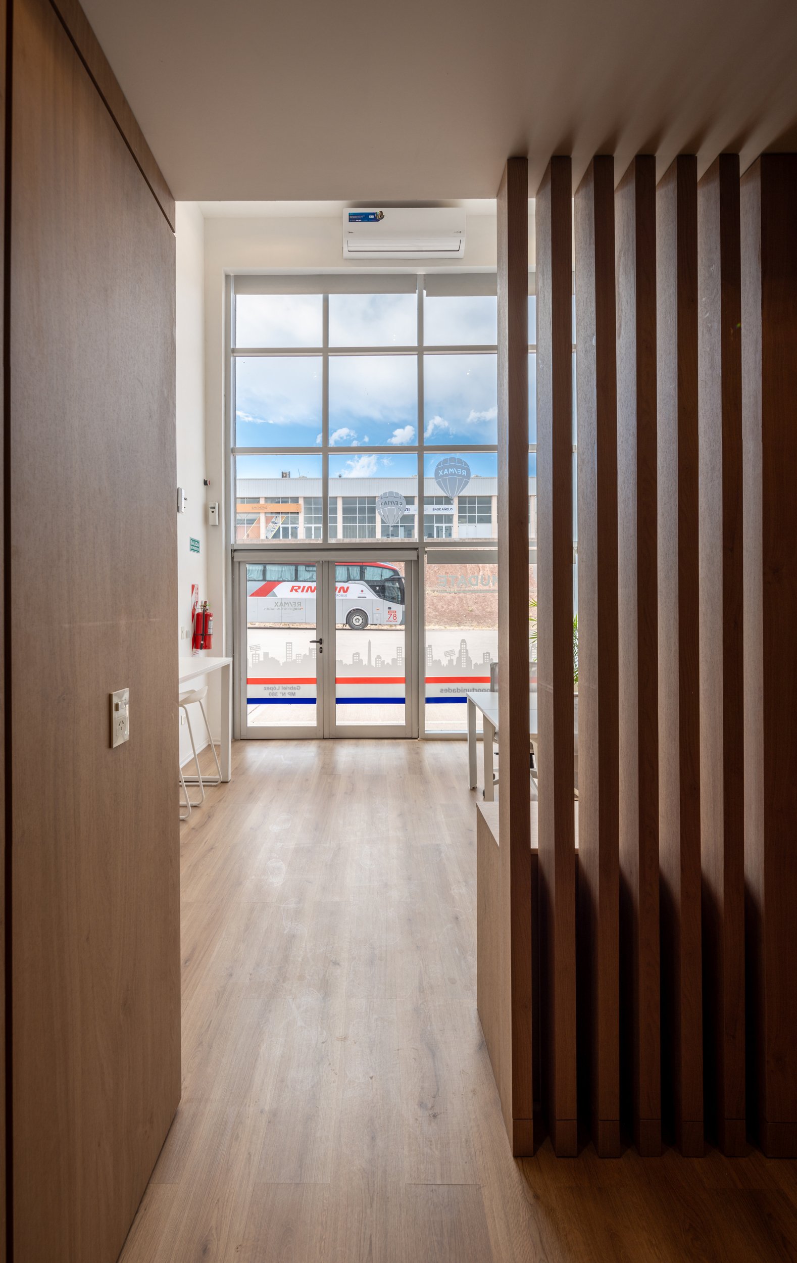 Interior de un espacio con piso de madera, una pared de madera en el lado izquierdo, una estructura de madera en el lado derecho, una ventana grande con cielo y edificios visibles, y una puerta de vidrio con logotipo y vehículos afuera.