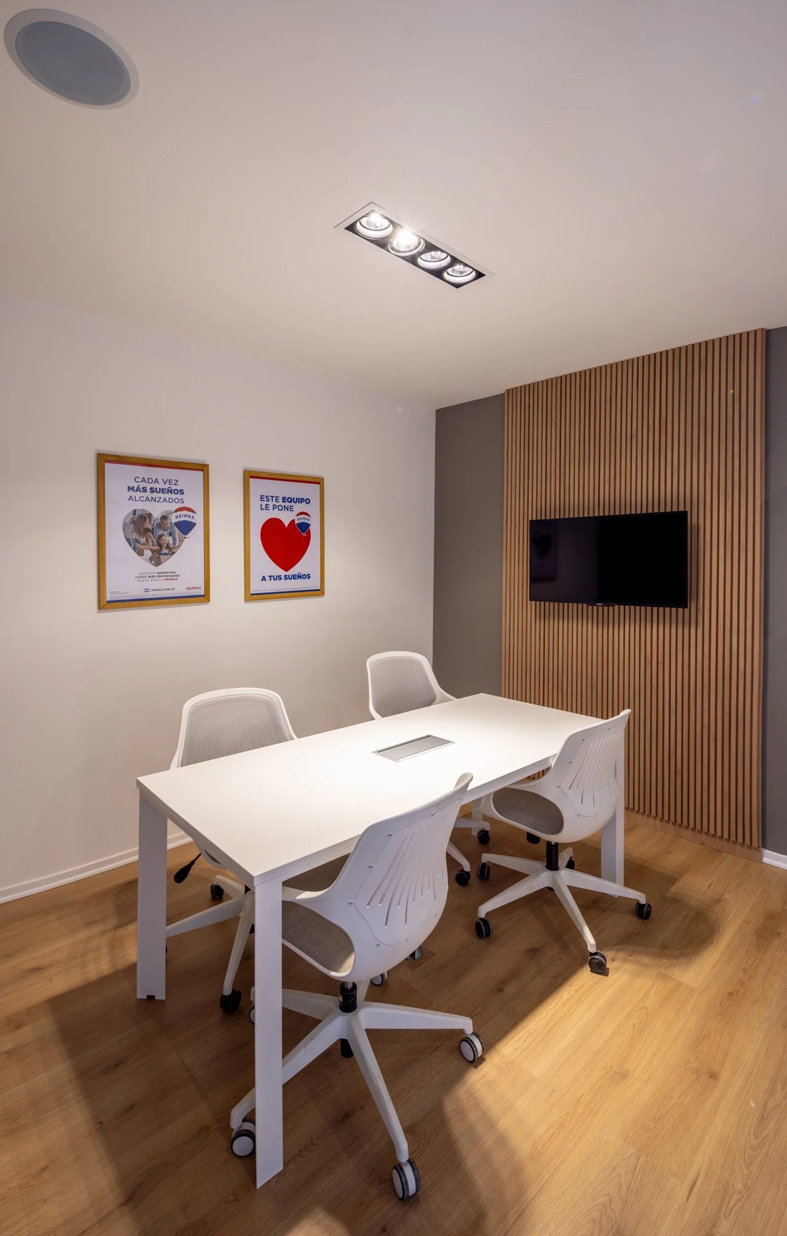 Sala de reuniones con mesa blanca, cuatro sillas blancas con ruedas, pared con panel de madera, televisión y dos carteles en la pared, iluminación en techo.
