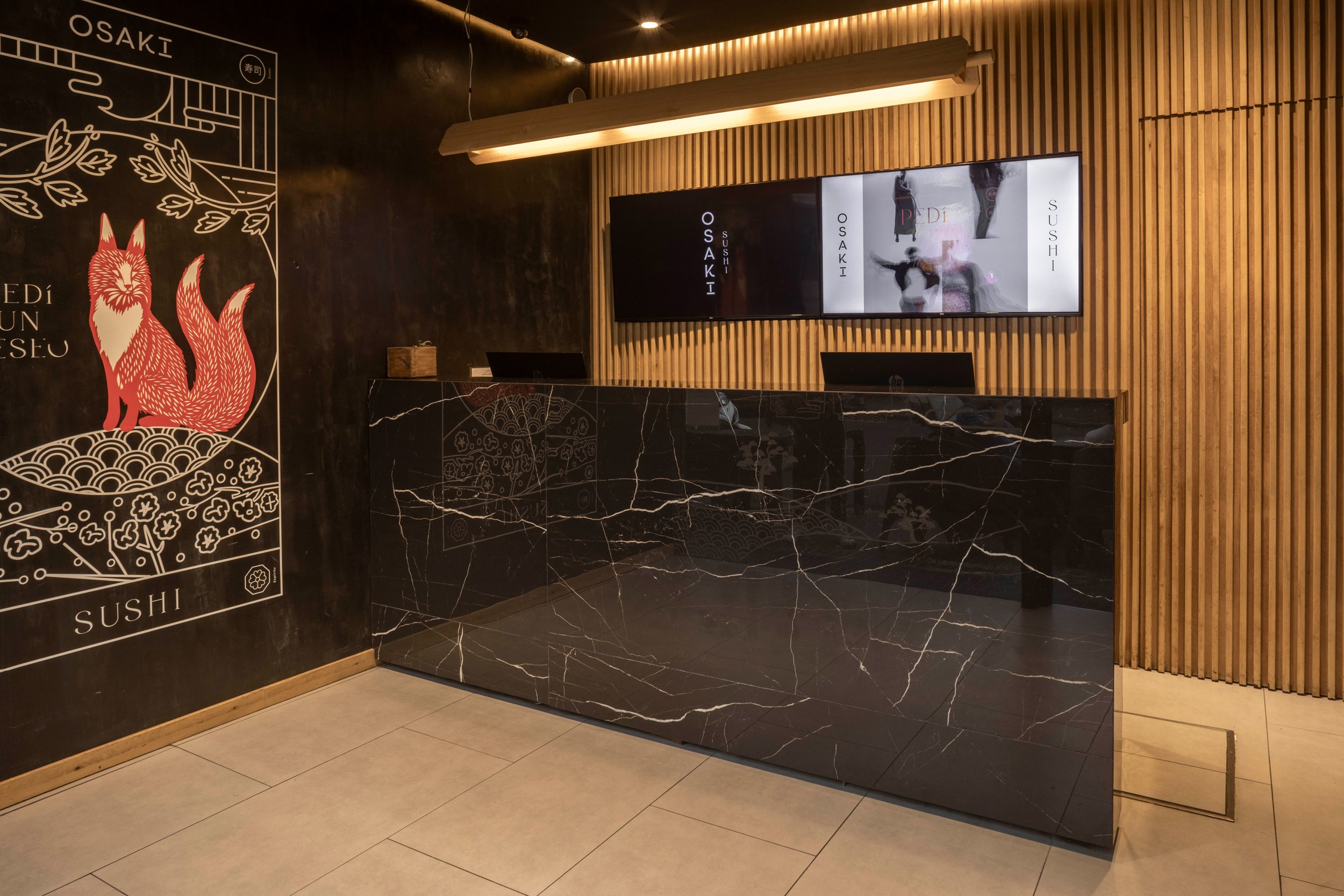Mostrador de recepción de un restaurante de sushi con paredes de madera y una pantalla digital. Decoración con un dibujo de un zorro en una pared negra.