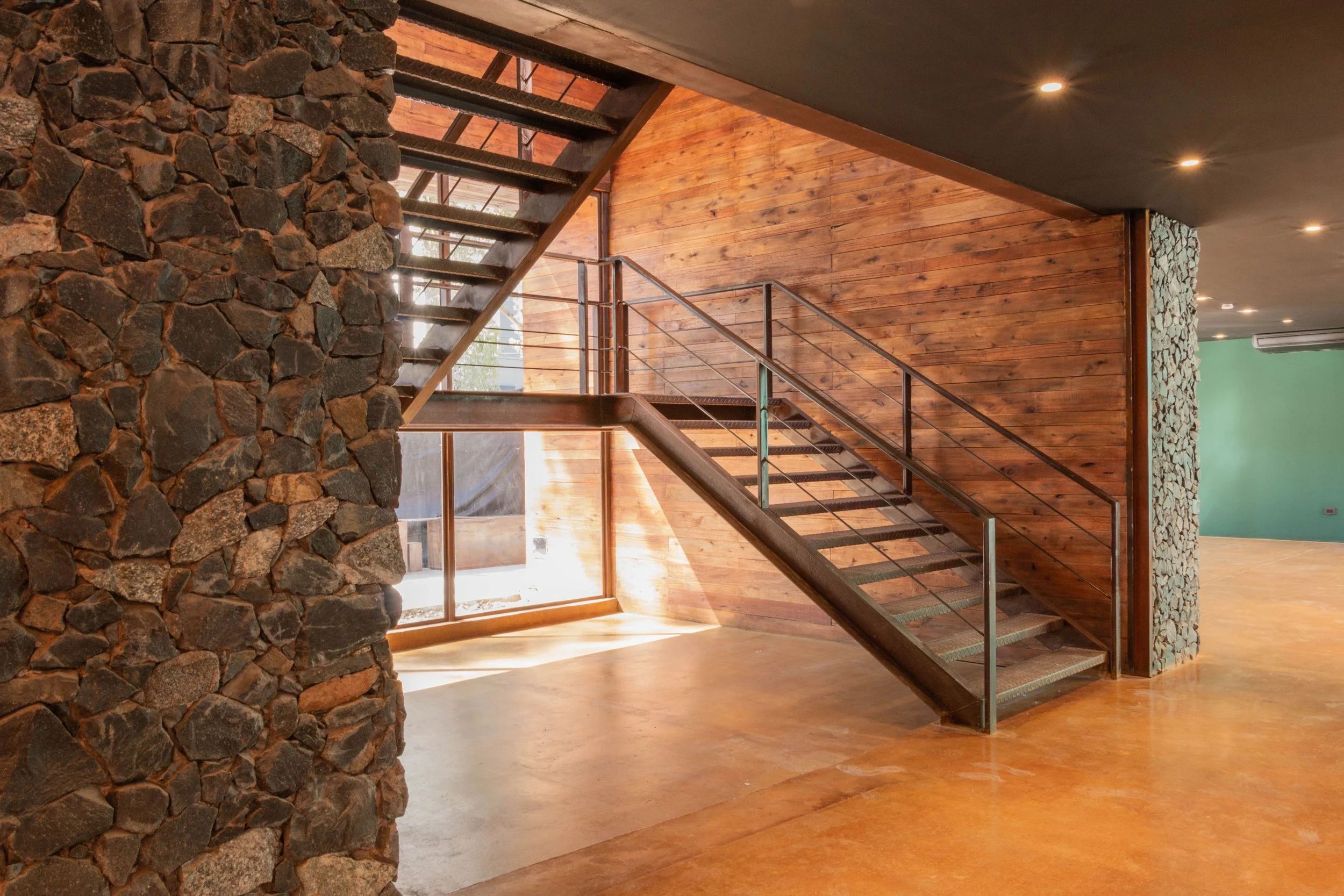 Interior moderno con escalera de metal y madera, paredes de piedra y madera, suelo de color cálido, y ventanas grandes que dejan entrar luz natural.