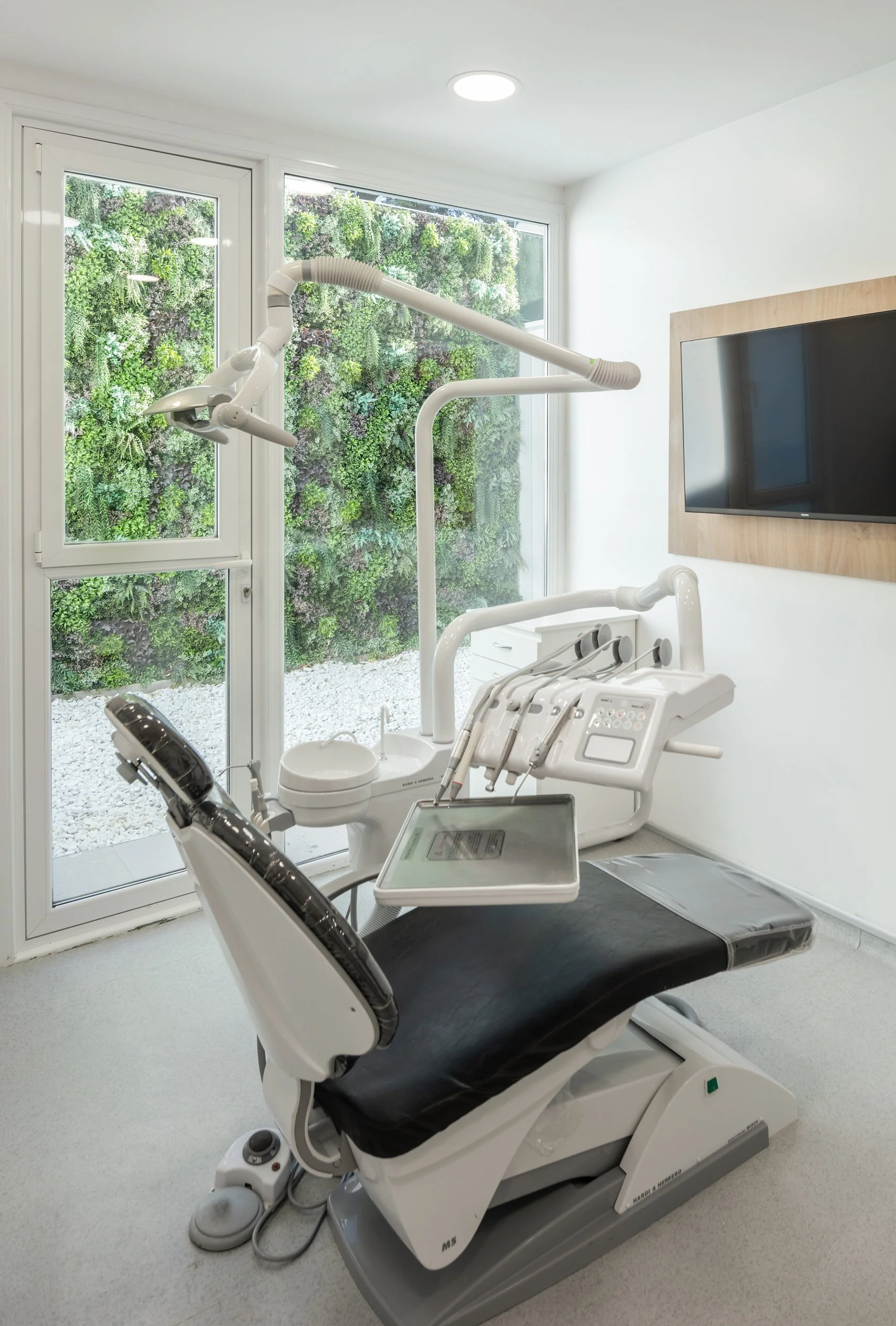 Consultorio dental con silla y equipo dental. Ventana grande con vista a un jardín vertical.