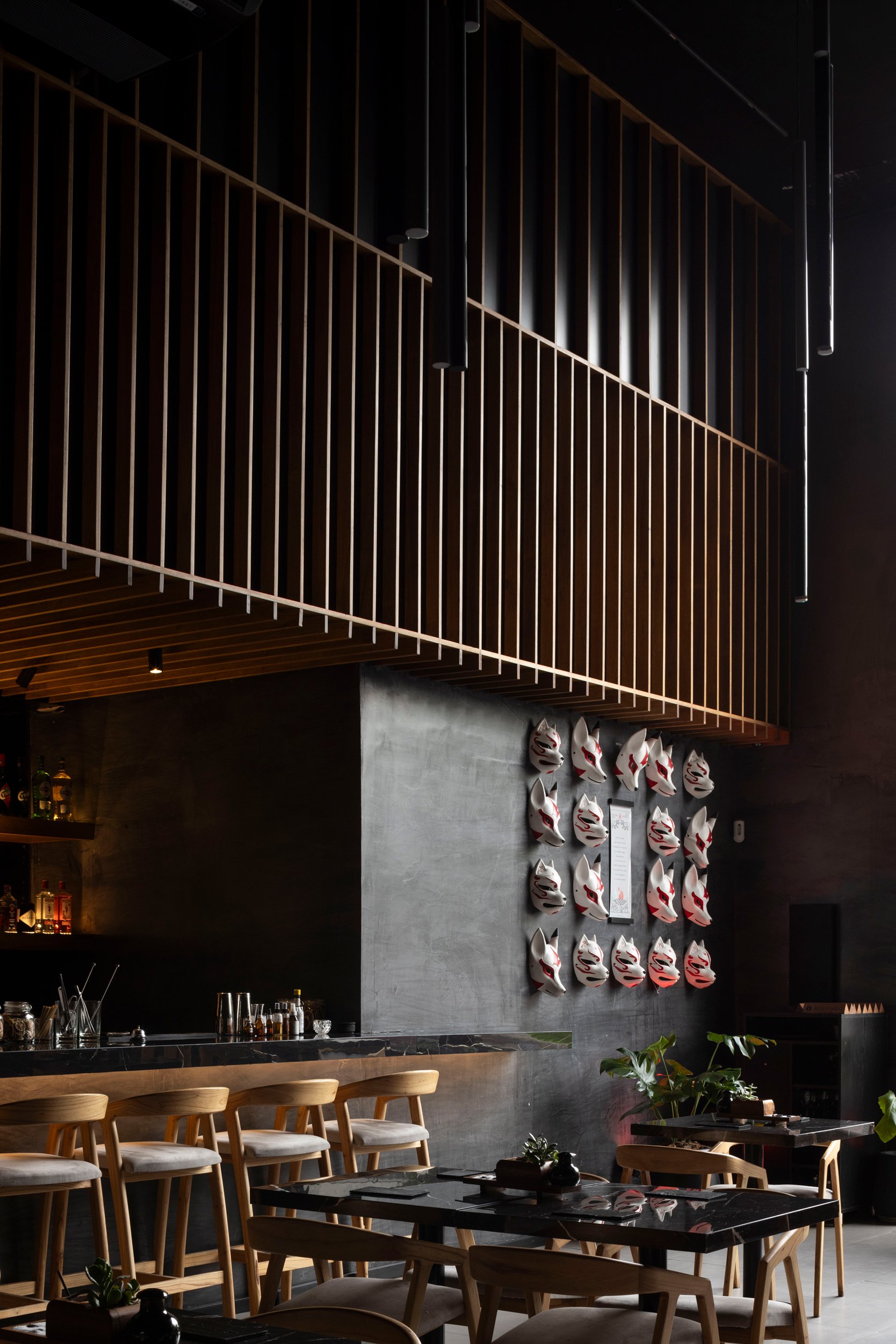 Interior de un restaurante con barra de madera, sillas de madera, una pared decorada con máscaras japonesas y plantas verdes