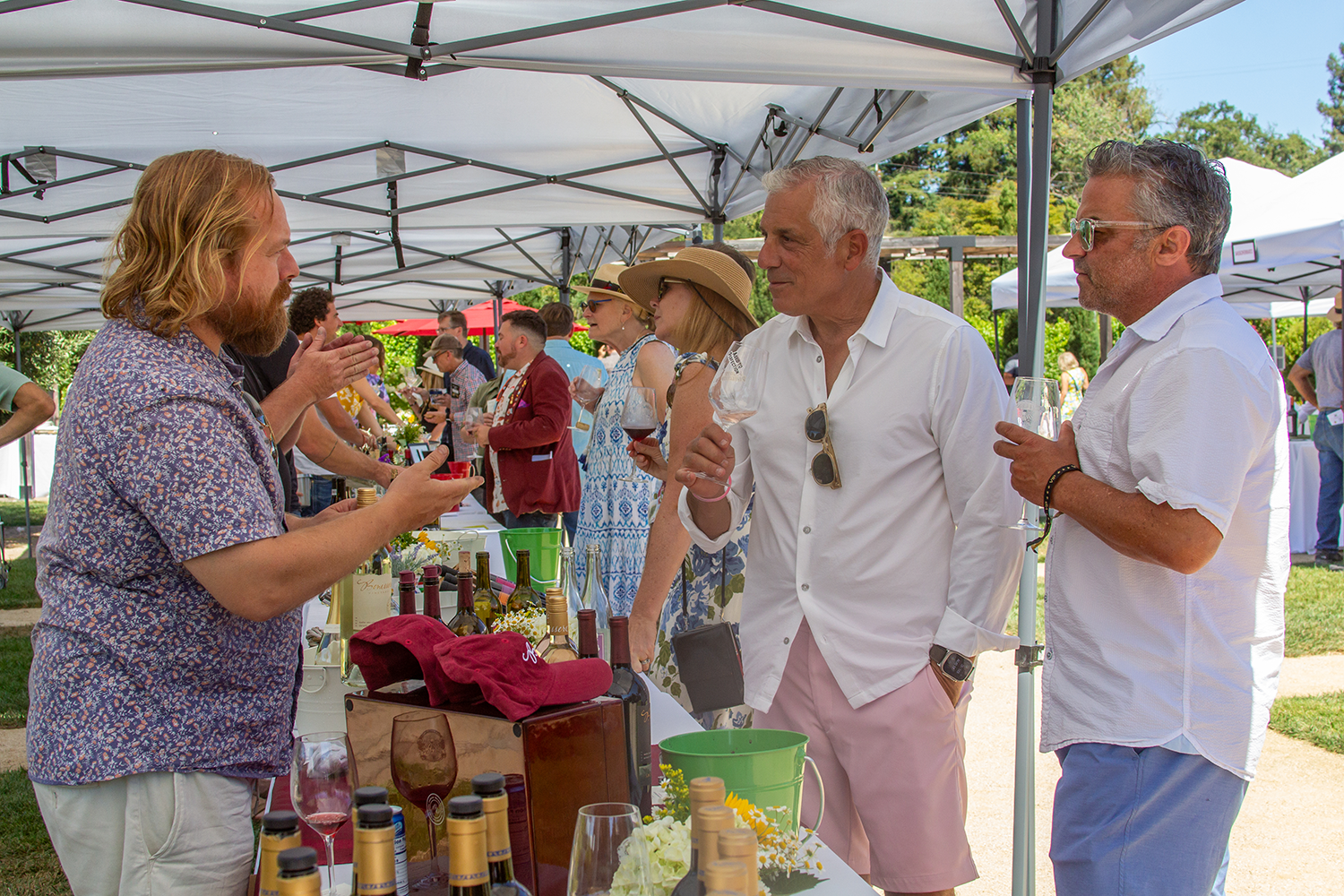People at an outdoor wine tasting event under white tents, examining bottles and glasses of wine.