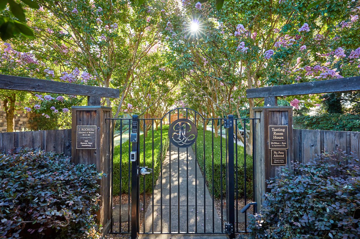 Gated entrance to a pathway surrounded by lush trees with purple flowers, sunlight shining through the branches, and signs indicating vineyard and winery information.